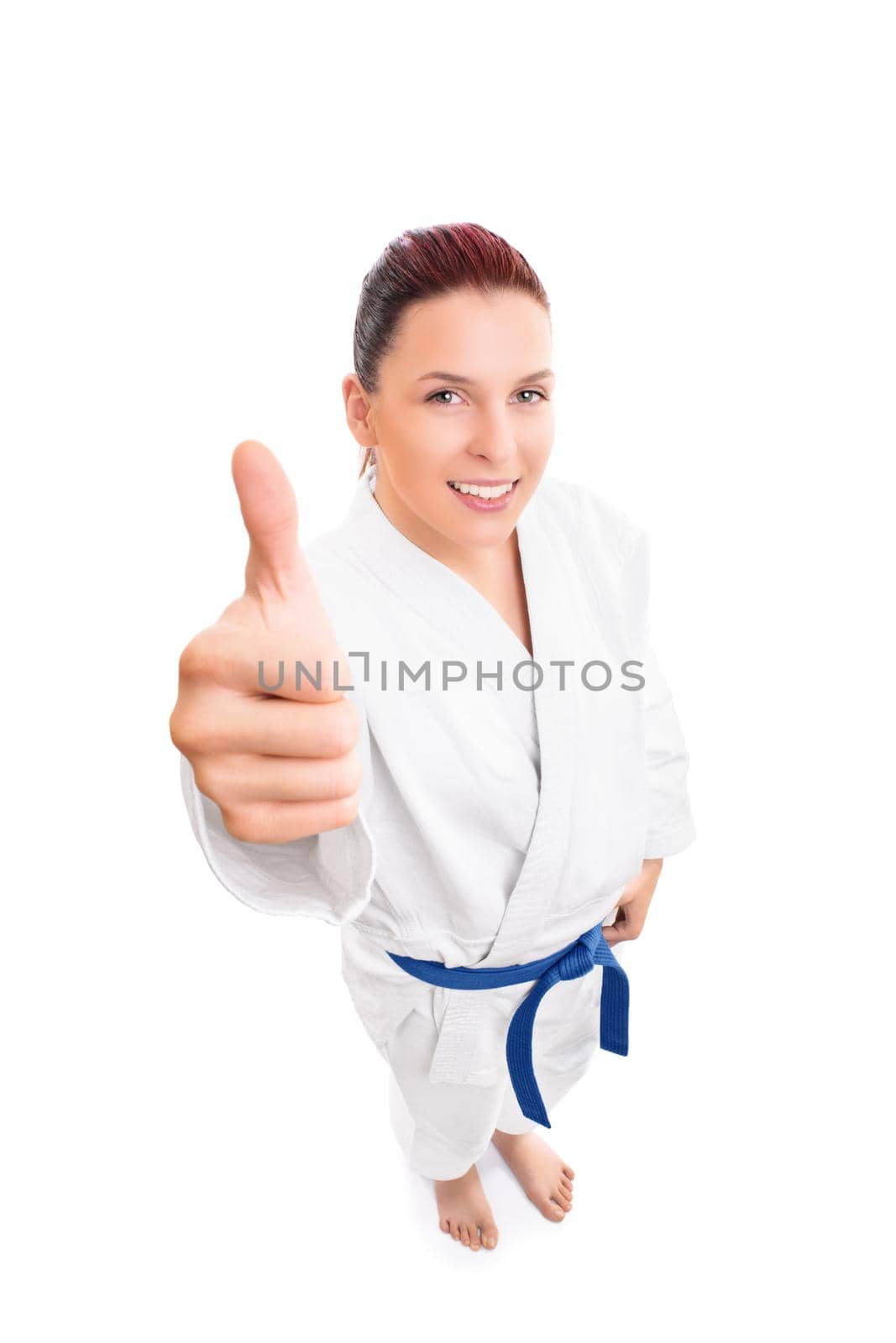 Top-down full body portrait of a beautiful smiling young female fighter in white kimono and blue belt showing thumb up gesture, isolated on white background. Martial arts concept.