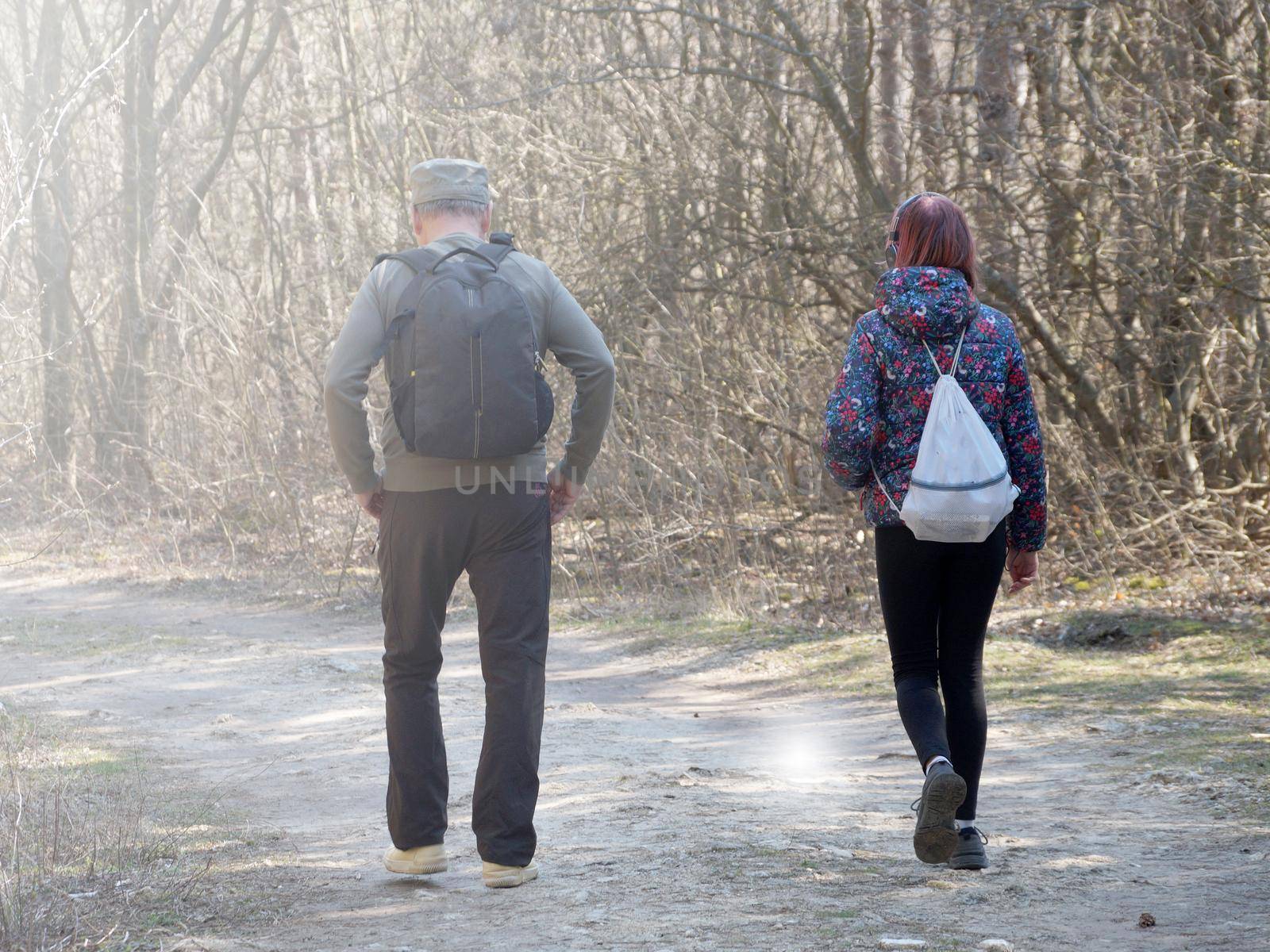a man and a teenage girl are walking with backpacks along a forest road on a sunny day, back view