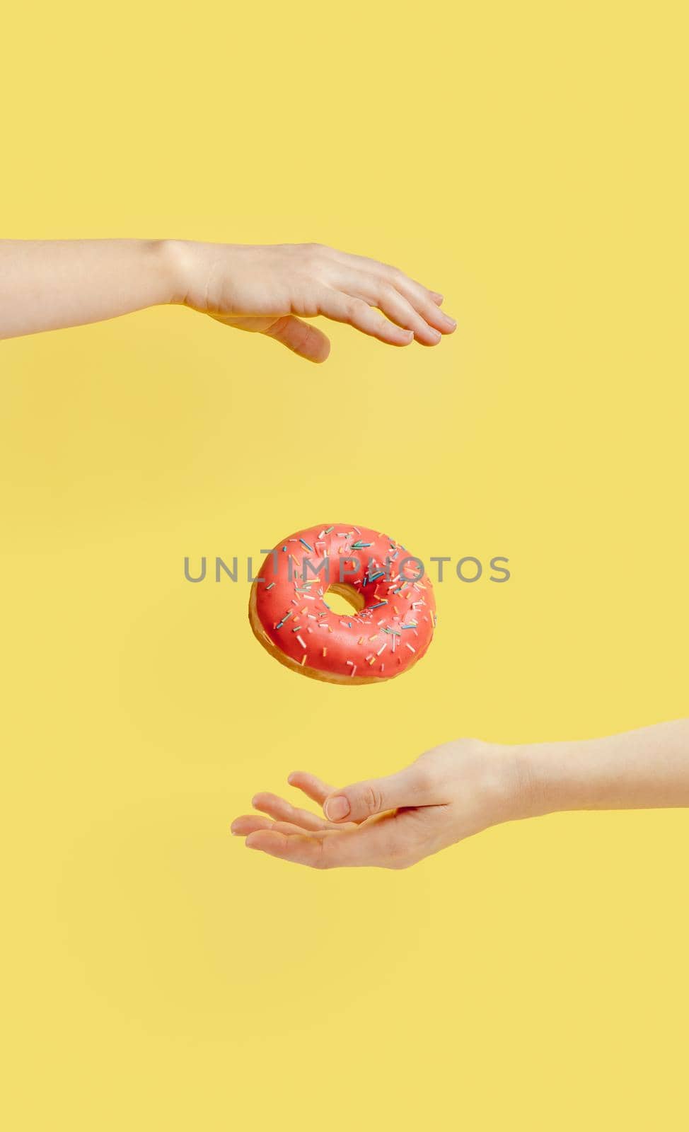 Donut falls between hands on bright yellow paper background. Donuts delivery, diet, sugar concept. Female hands and glazed donut with sprinkles flying in air. Vertical. Creative still life