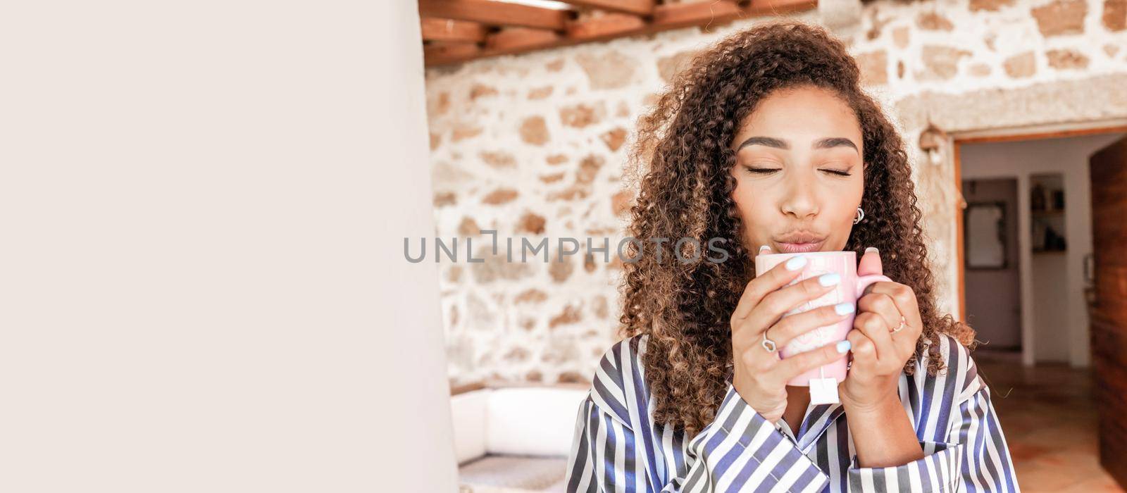 Beautiful black Hispanic girl with dark curly hair in striped silk pajamas enjoying her cup of hot tea holding it in her hands with her eyes closed on the porch of her country house. Left copy space