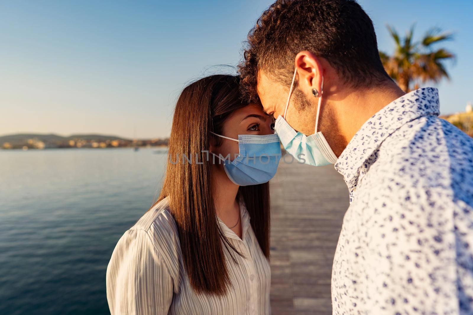 Man and woman look into each other's eyes by leaning foreheads against each other wearing protective mask due to Coronavirus. Young couple of tourists at sunset or dawn in ocean resort with pandemic