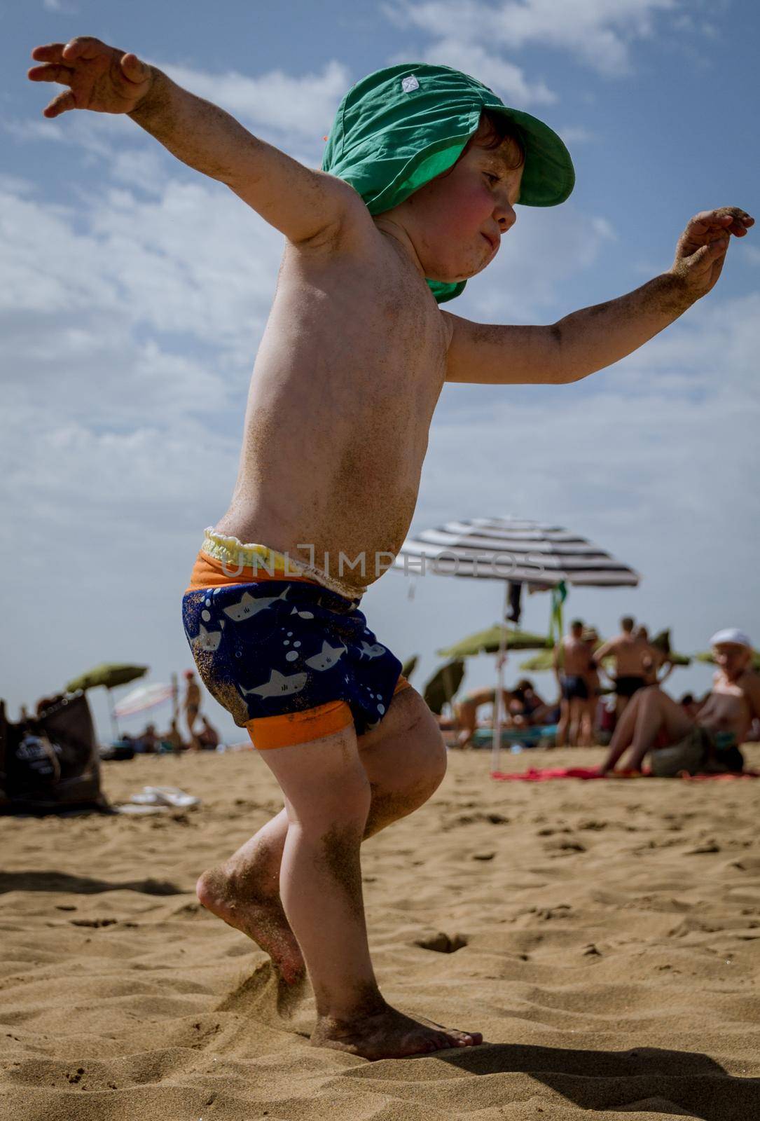 A cute, red head baby boy wearing a green hat and swimming suit walking and playing in the sand