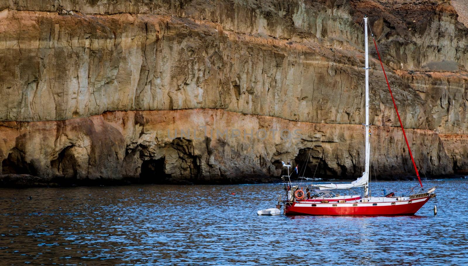 Wide shot of a red boat floating in the sea with a rocky formation in the background