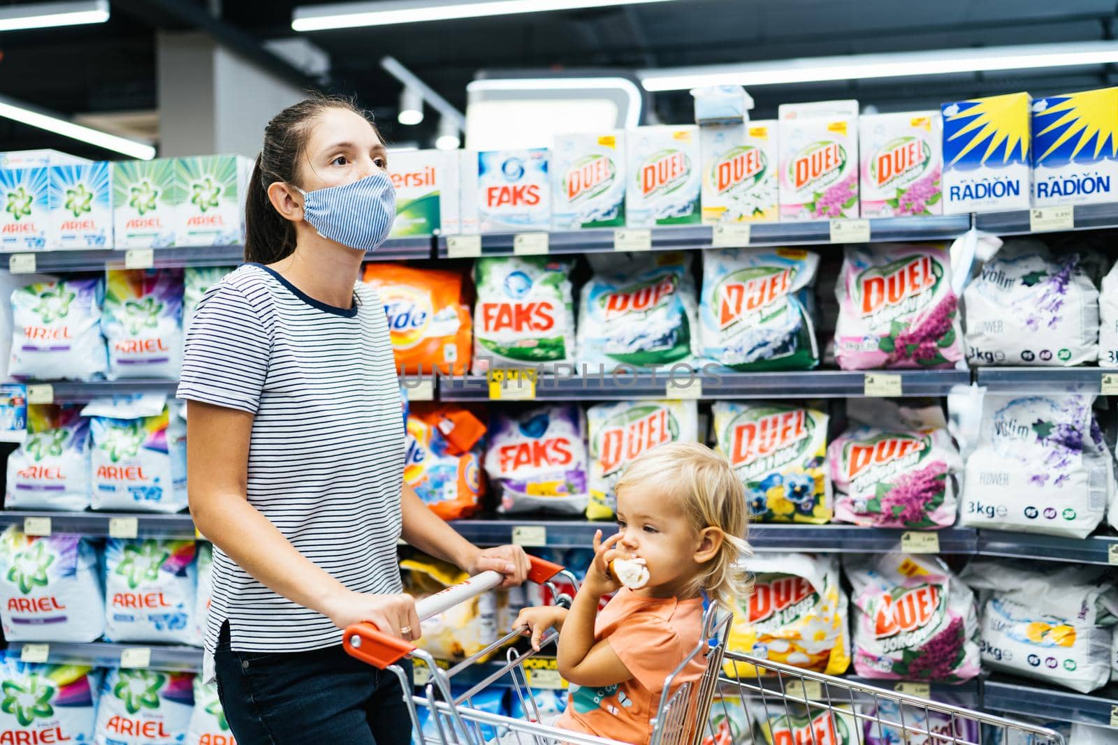 Budva, Montenegro - 17 march 2021: Shopping with children during the coronavirus epidemic. Mother and baby in protective mask buying fruit in the supermarket. Mom and little girl household chemicals by Nadtochiy