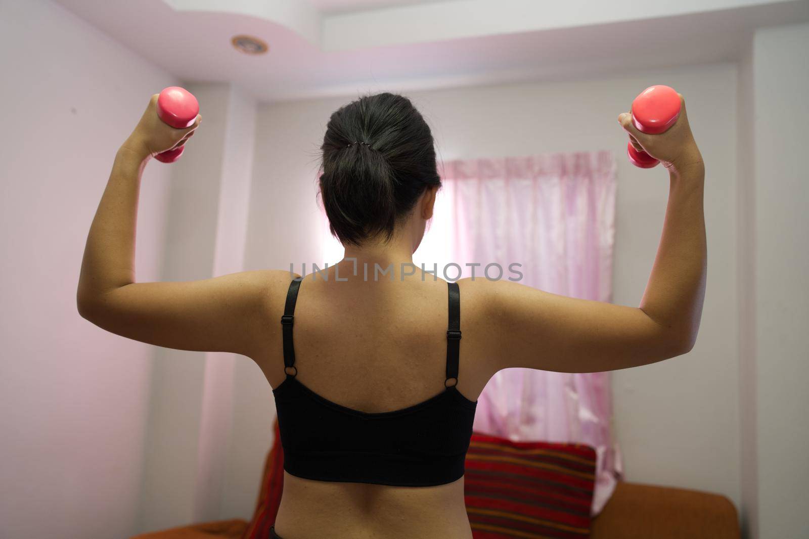 Asian women training to lift pink dumbbells for building muscle, the concept of staying healthy by weight loss and recreation at their residence. by noppha80