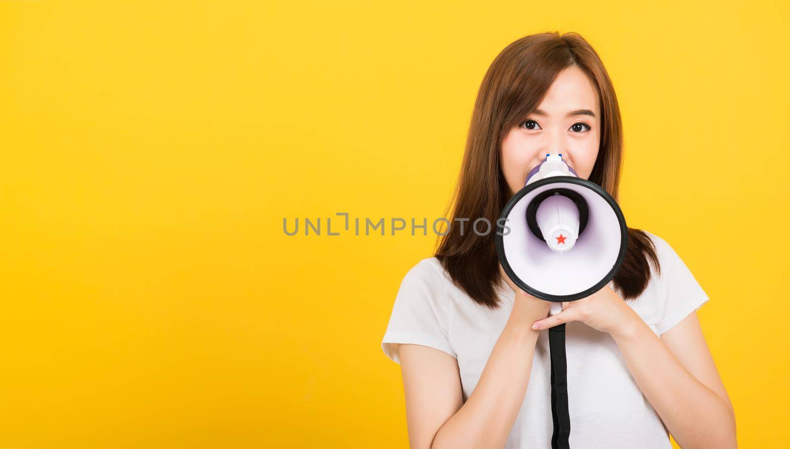 Asian happy portrait beautiful cute young woman teen standing making announcement message shouting screaming in megaphone looking to camera isolated, studio shot on yellow background with copy space