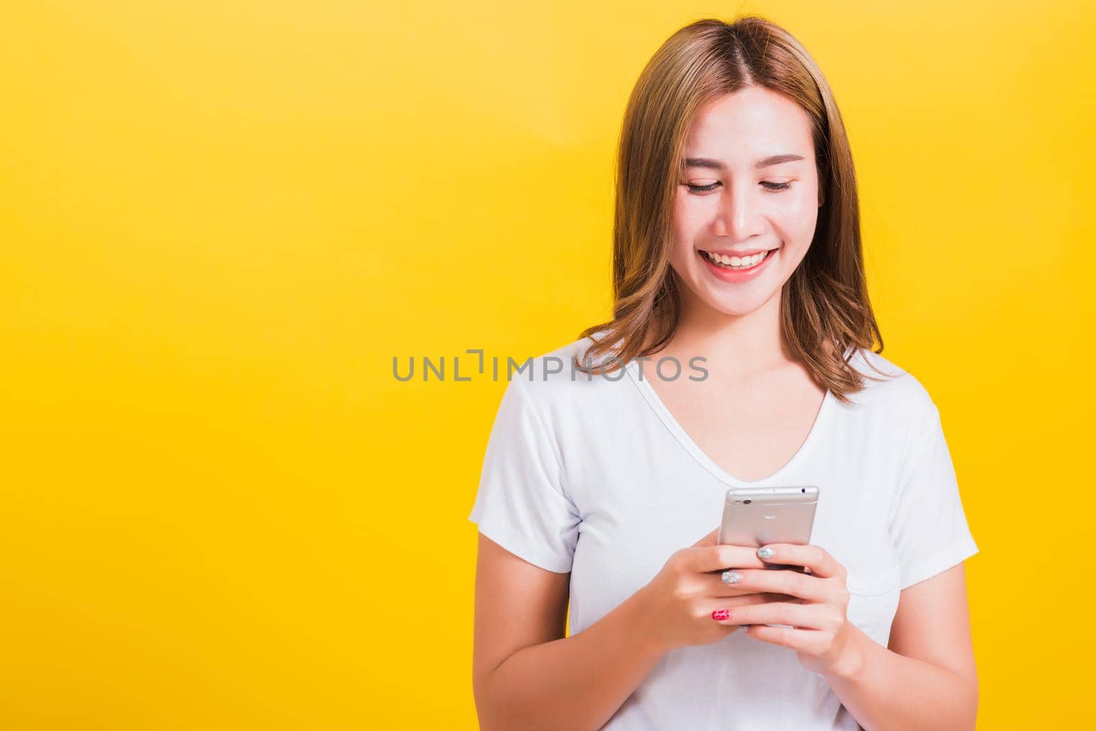 Asian Thai portrait happy beautiful cute young woman smile standing playing game or writing SMS message on smartphone looking to the phone, studio shot isolated on yellow background with copy space