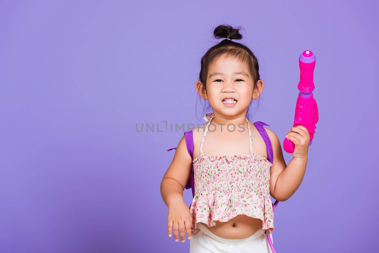 Thai child funny hold toy water pistol and smile, Happy Asian little girl holding plastic water gun, studio shot isolated on purple background, Thailand Songkran festival day national culture concept