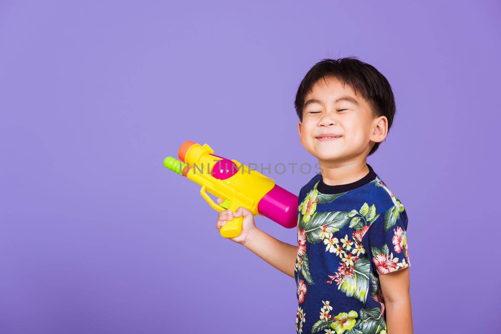 Thai kid funny hold toy water pistol and smiling, Happy Asian little boy holding plastic water gun, studio shot isolated on purple background, Thailand Songkran festival day national culture concept