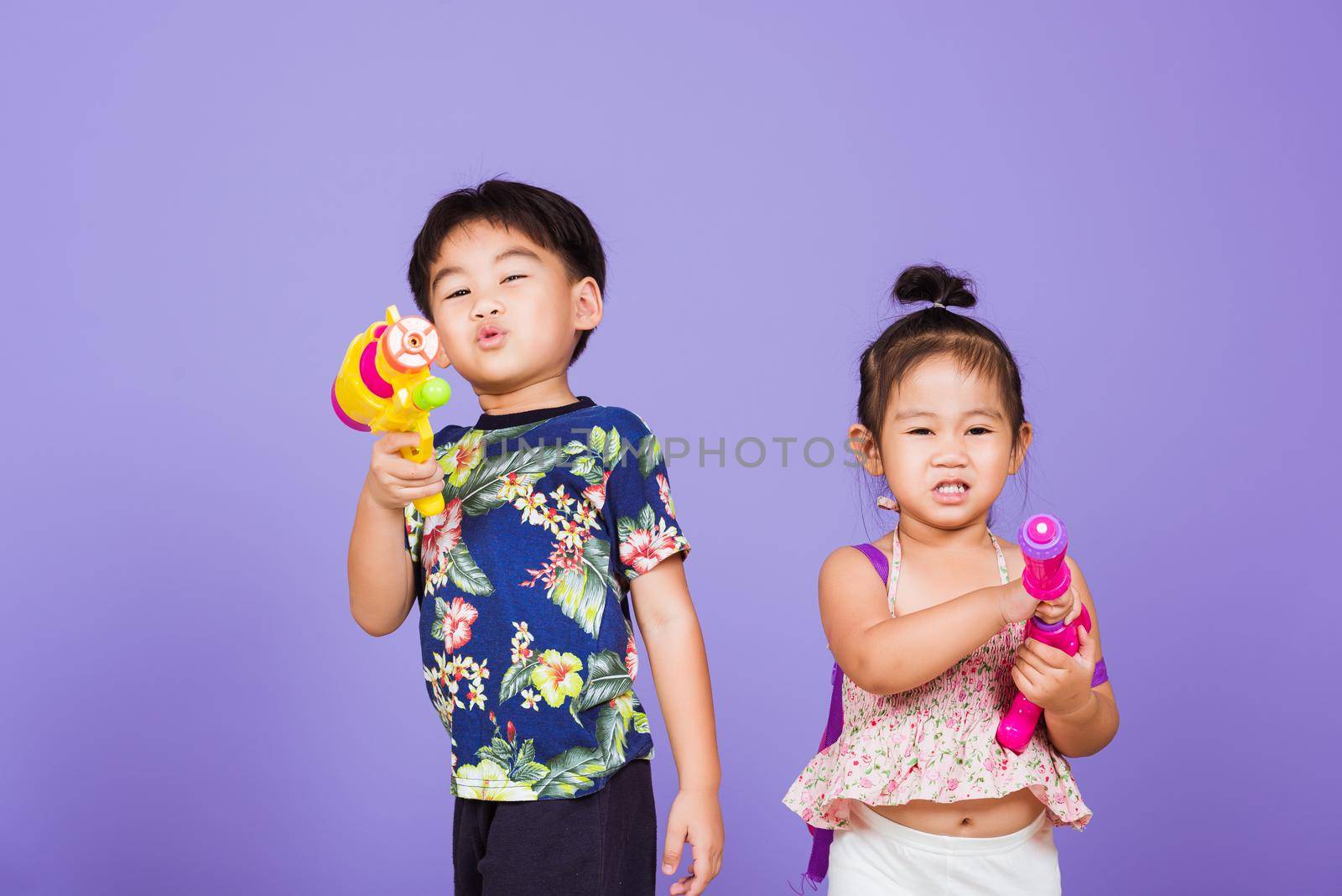 Two Happy Asian little boy and girl holding plastic water gun, Thai children funny hold toy water pistol and smile, studio shot isolated on purple background, Thailand Songkran festival day culture.