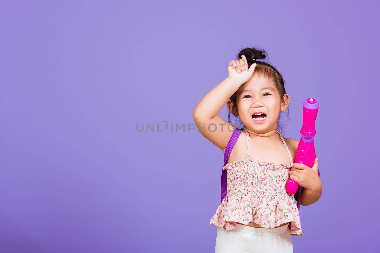 Happy Asian little girl holding plastic water gun, Thai child funny hold toy water pistol and smile, studio shot isolated on purple background, Thailand Songkran festival day national culture concept