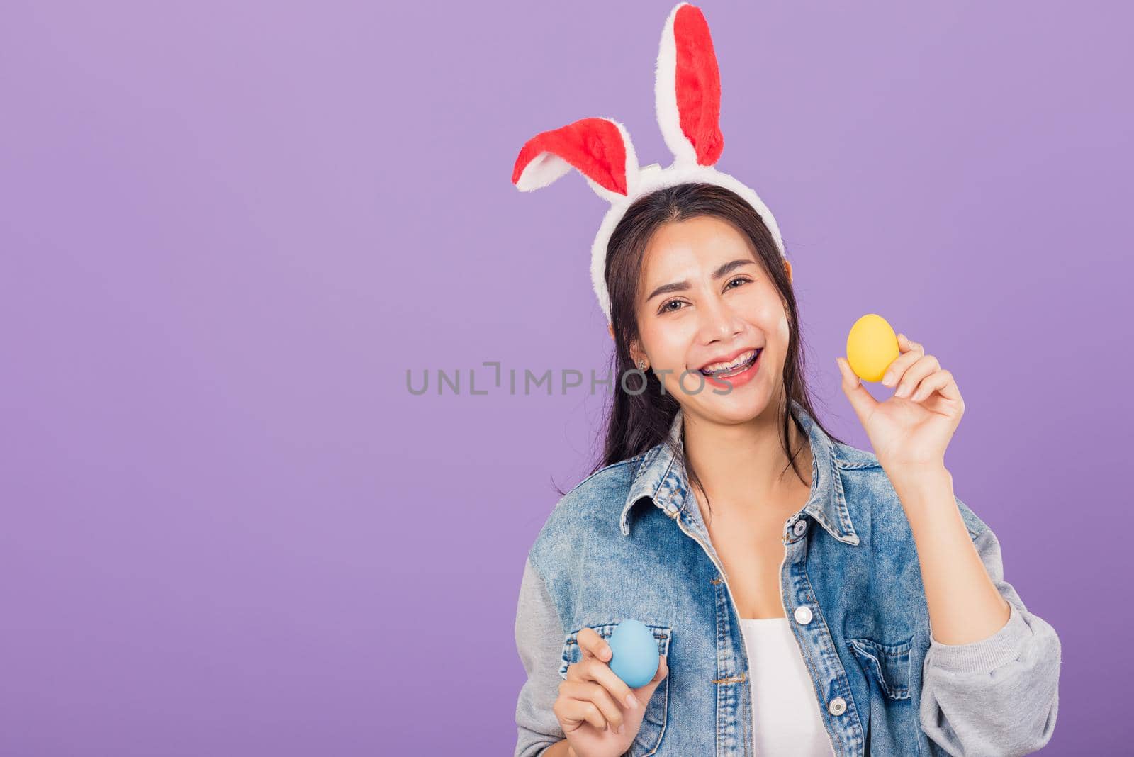 Happy Easter concept. Beautiful young woman smiling wearing rabbit ears and denims hold colorful Easter eggs gift on hands, Portrait female looking at camera, studio shot isolated on purple background