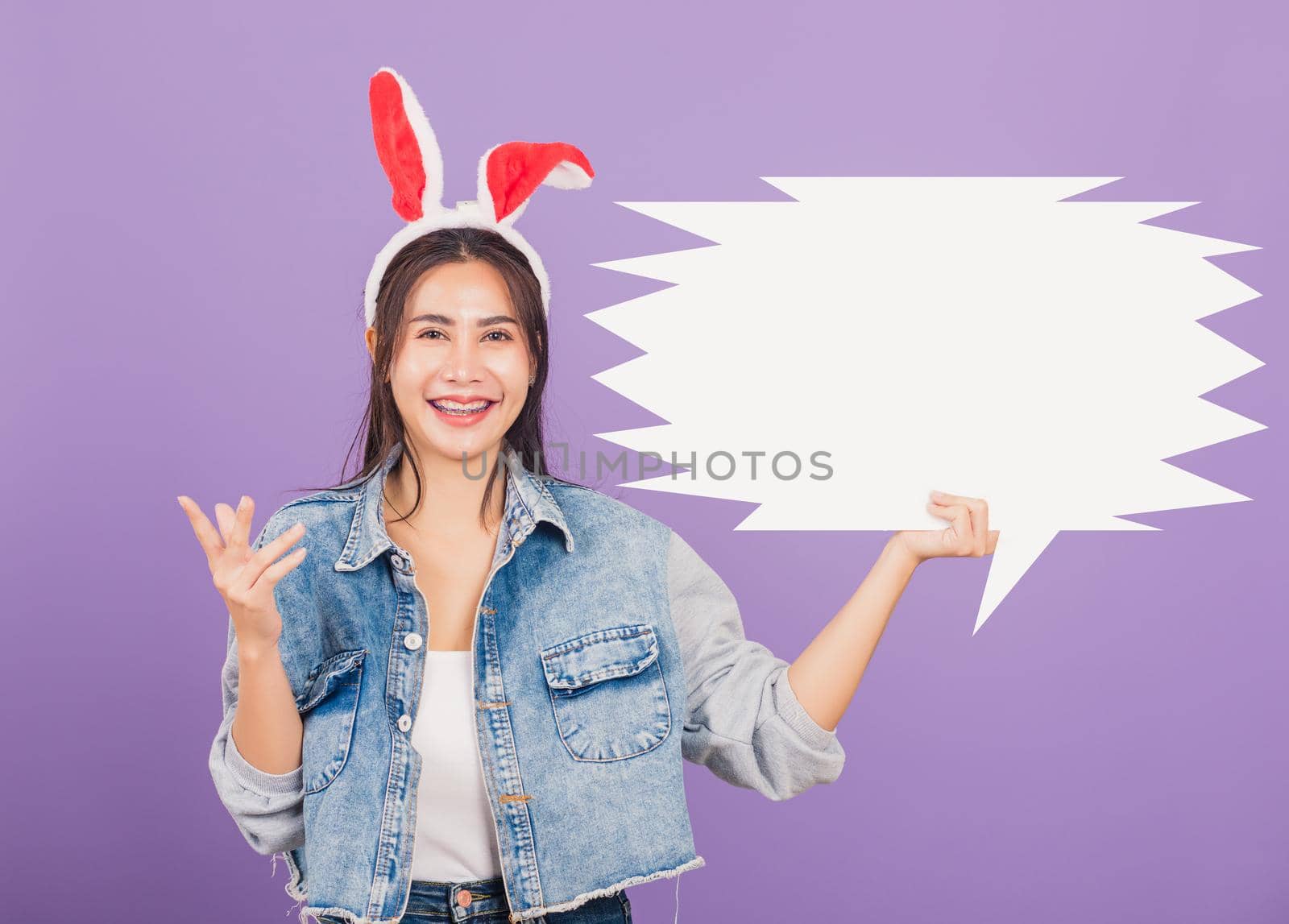 
Happy Easter Day. Beautiful young woman smiling excited wearing rabbit ears and denims holding empty speech bubble, Portrait female looking at camera, studio shot isolated on purple background