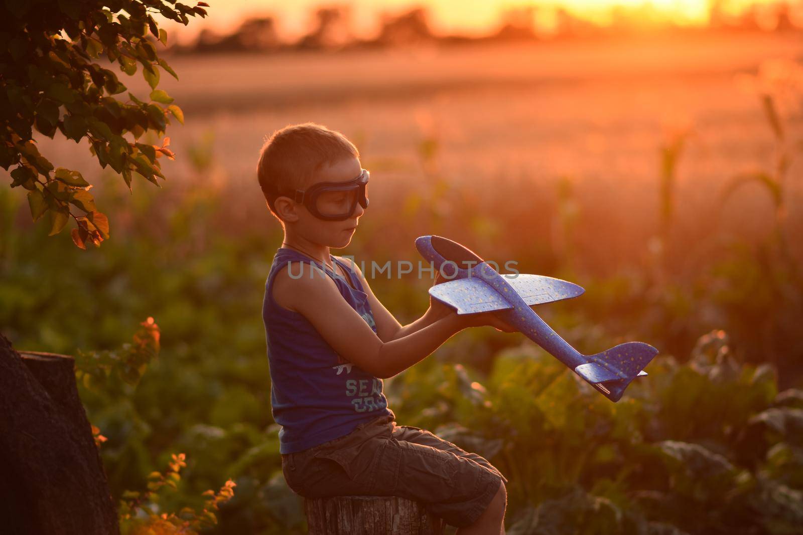 A boy with pilot's glasses is playing with an airplane. The child imagines himself as an aviator pilot. Outdoor games in summer at sunset.