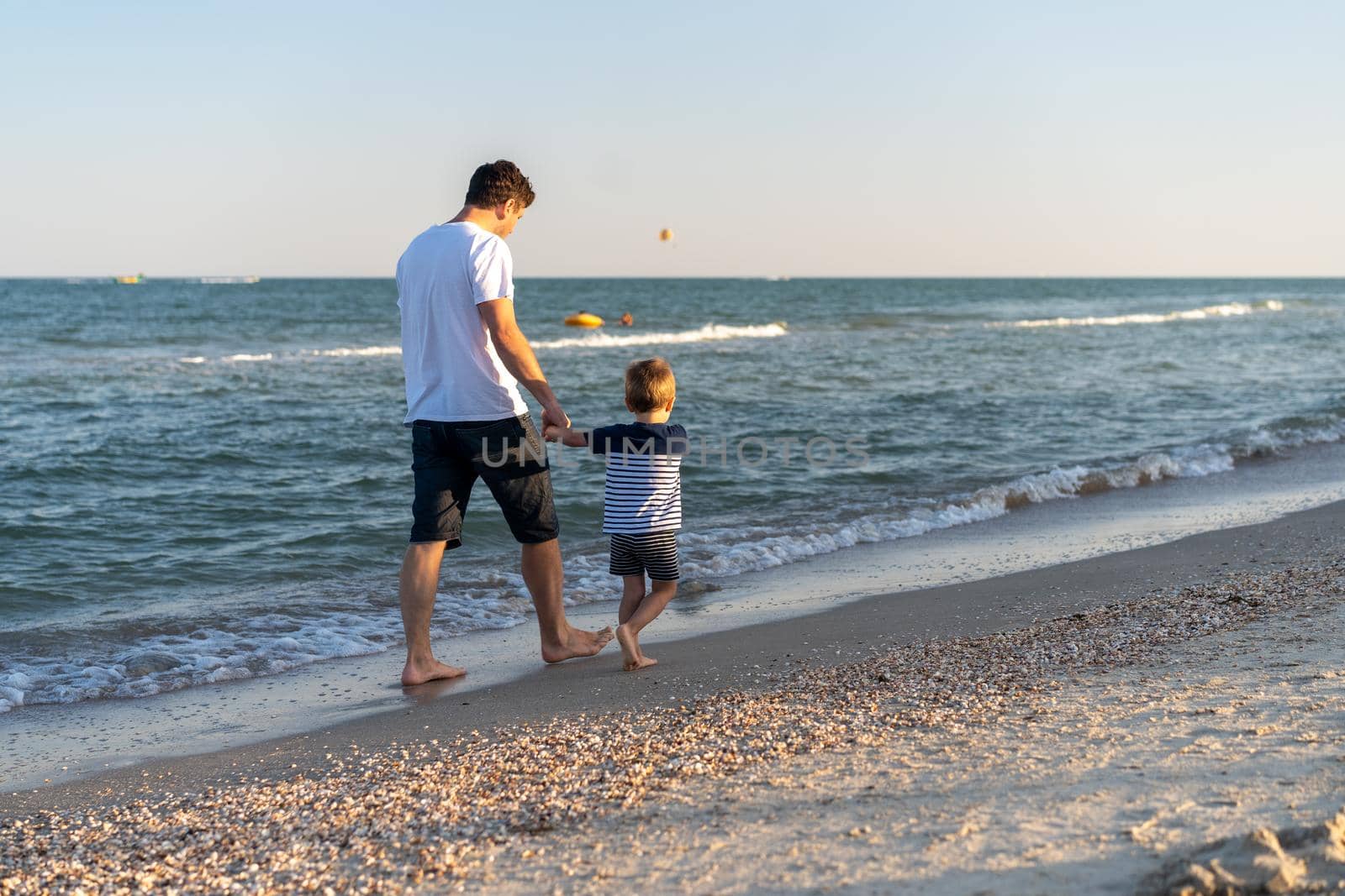 Young Caucasian dad with little son walk warm summer day along the sea coast. Summer family vacation concept. Friendship father and son. Happy childhood on the beach with parent. free space for text