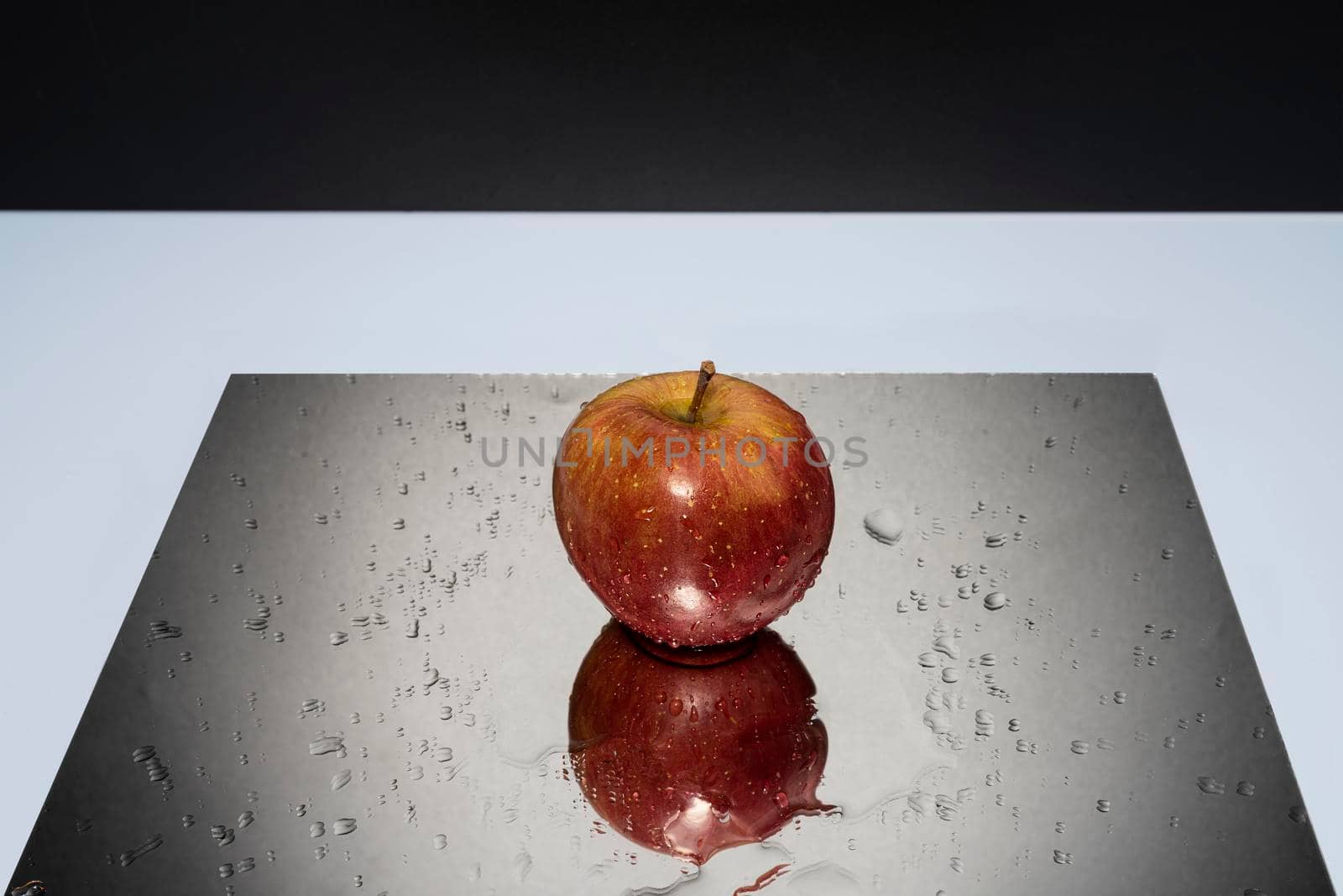 A red apple resting on a mirror with falling water drops