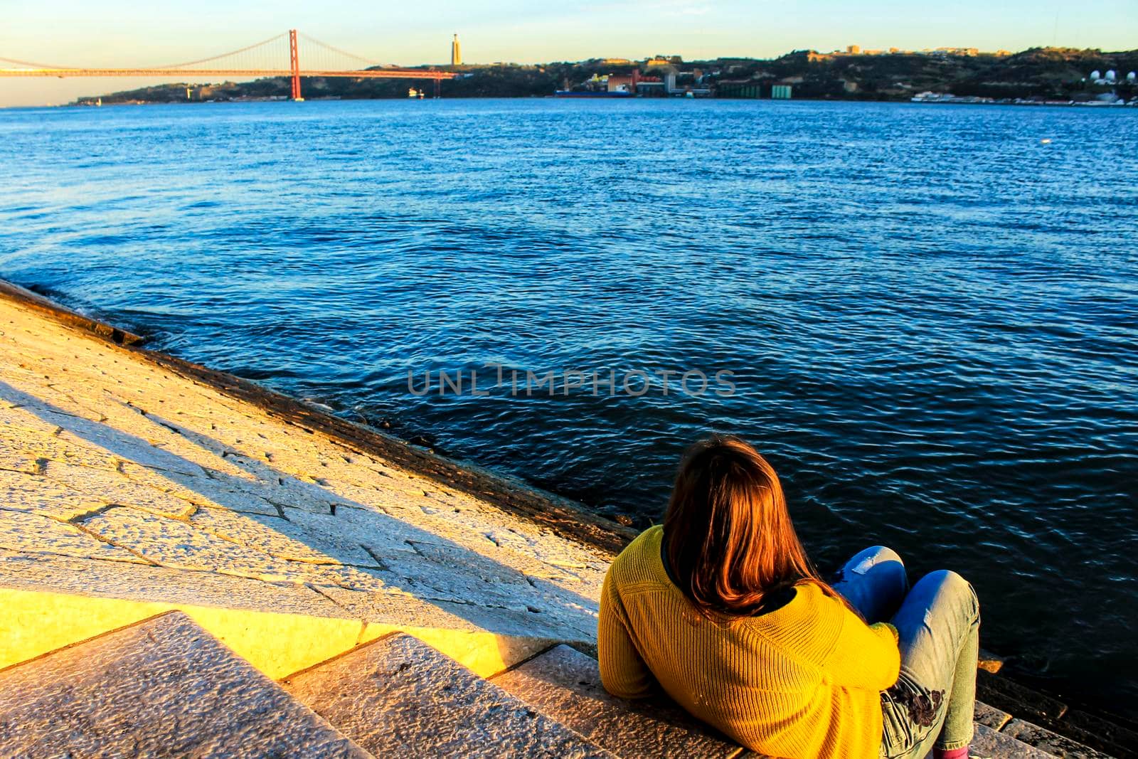 Girl sitting next to the riverbank at sunset in Lisbon by soniabonet