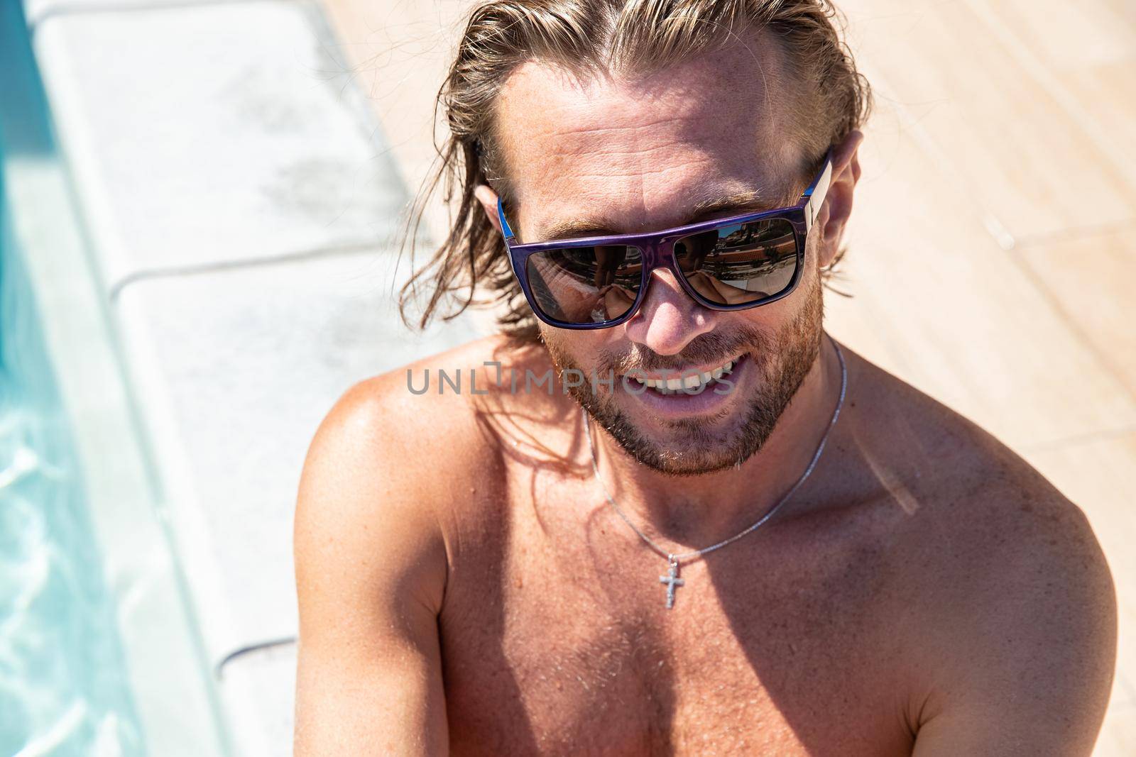 The handsome brutal man in sunglasses with a long hair and naked torso sits near the pool, a sports suntanned body, sunglasses with a blue frame, he is red blue swimming shorts, sunny day