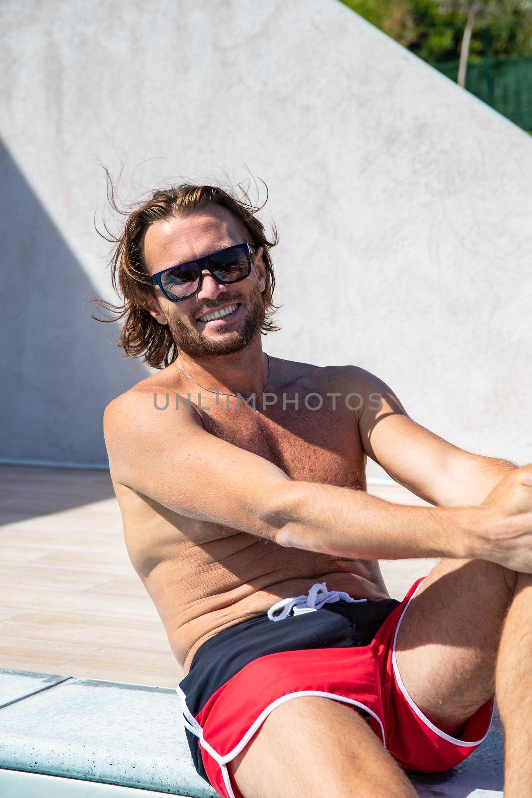 The handsome brutal man in sunglasses with a long hair and naked torso sits near the pool, is looking a camera, big smile, a sports suntanned body, sunglasses with a blue frame, sunny