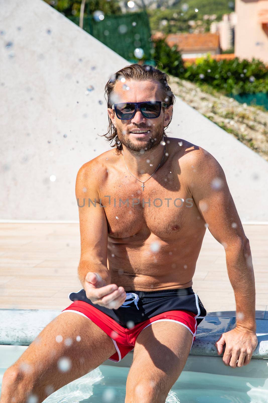 The handsome brutal man in sunglasses with a long hair and naked torso sits near the pool, a sports suntanned body, sunglasses with a blue frame, he is red blue swimming shorts, sunny day