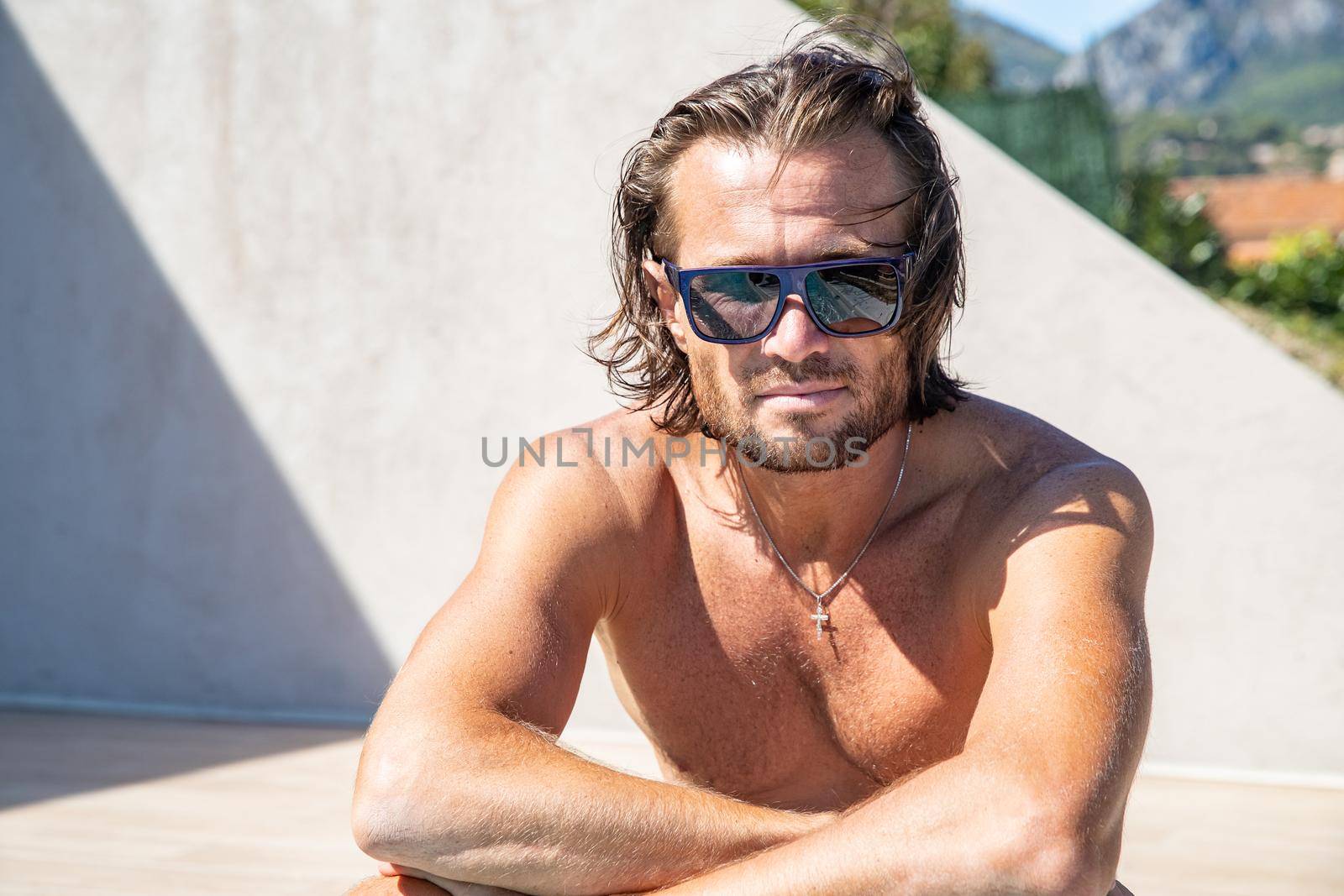 The handsome brutal man in sunglasses with a long hair and naked torso sits near the pool, a sports suntanned body, sunglasses with a blue frame, he is red blue swimming shorts, sunny day