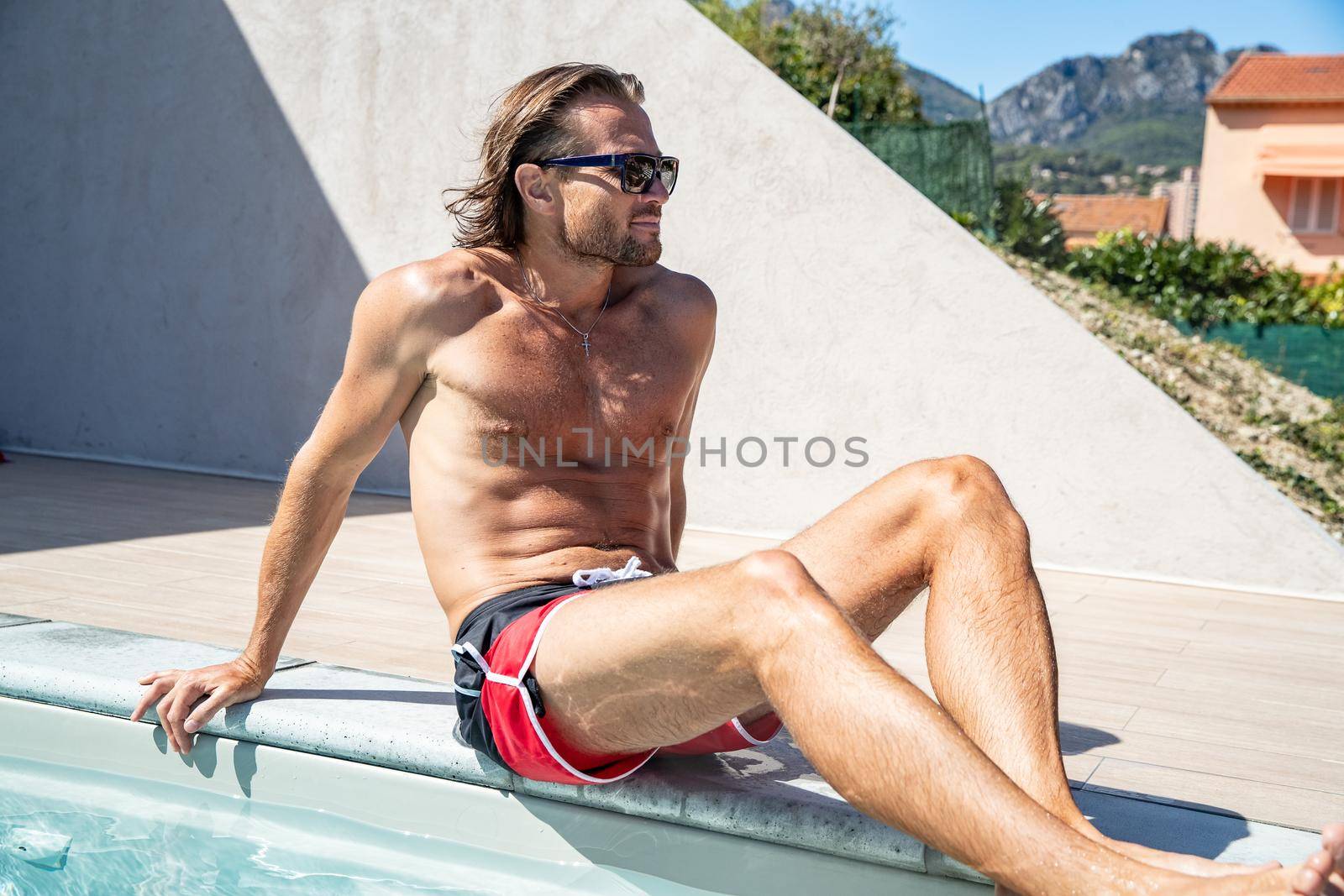 The handsome brutal man in sunglasses with a long hair and naked torso sits near the pool, a sports suntanned body, sunglasses with a blue frame, he is red blue swimming shorts, sunny day