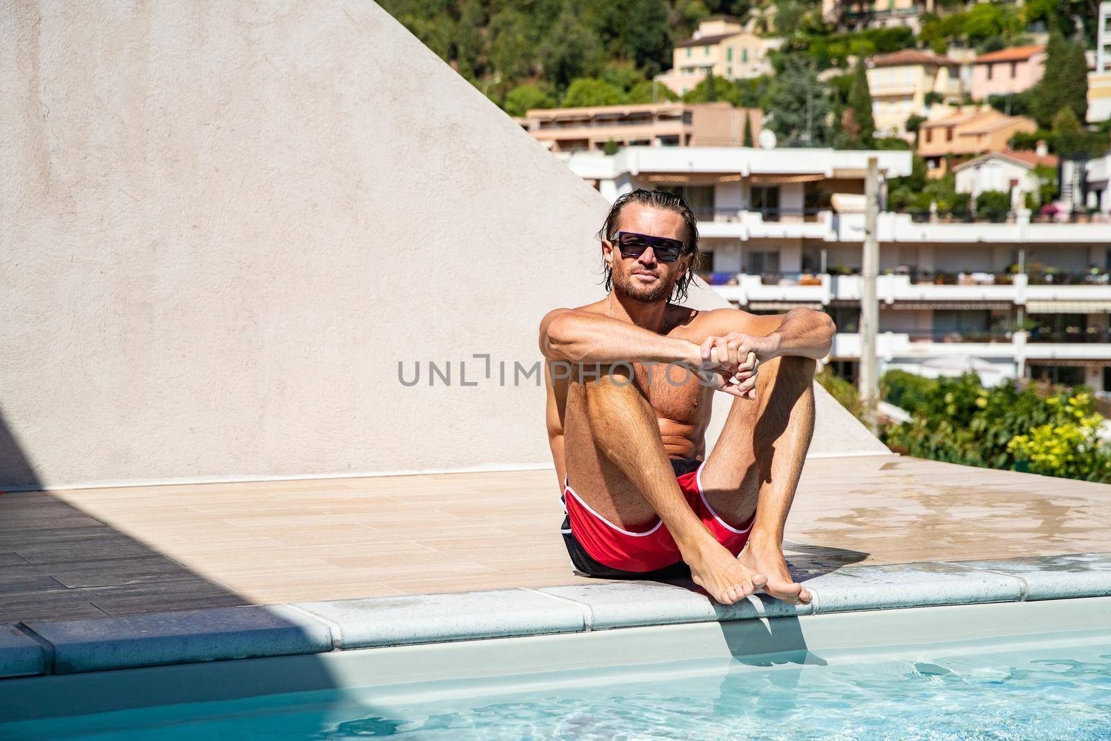 The handsome brutal man in sunglasses with a long hair and naked torso sits near the pool, a sports suntanned body, sunglasses with a blue frame, he is red blue swimming shorts, sunny day