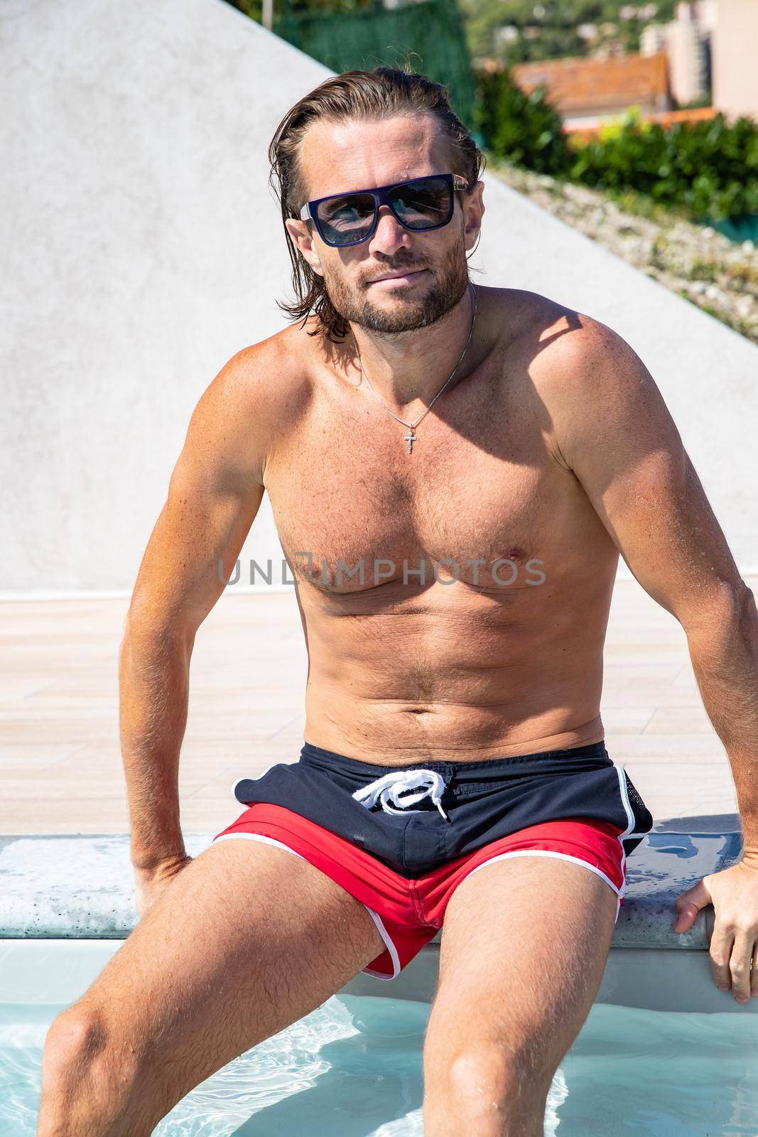 The handsome brutal man in sunglasses with a long hair and naked torso sits near the pool, a sports suntanned body, sunglasses with a blue frame, he is red blue swimming shorts, sunny day