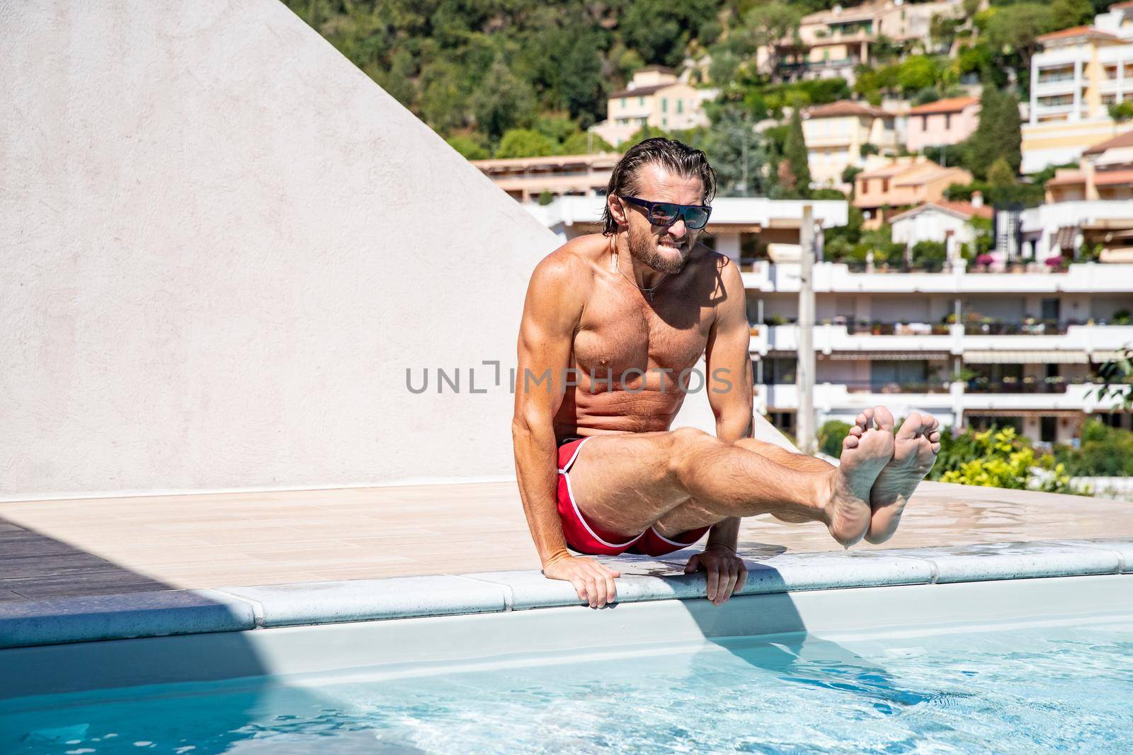 The handsome brutal man in sunglasses with a long hair and naked torso sits near the pool, a sports suntanned body, sunglasses with a blue frame, he is red blue swimming shorts, sunny day