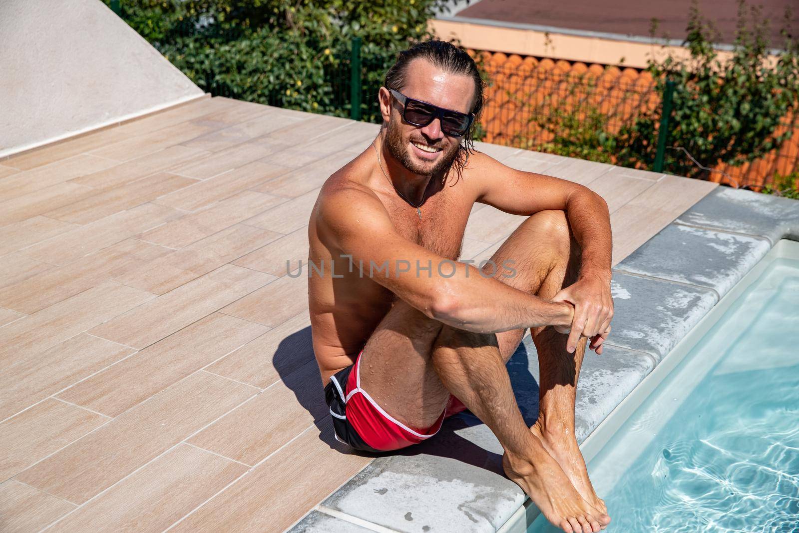 The handsome brutal man in sunglasses with a long hair and naked torso sits near the pool, a sports suntanned body, sunglasses with a blue frame, he is red blue swimming shorts, sunny day