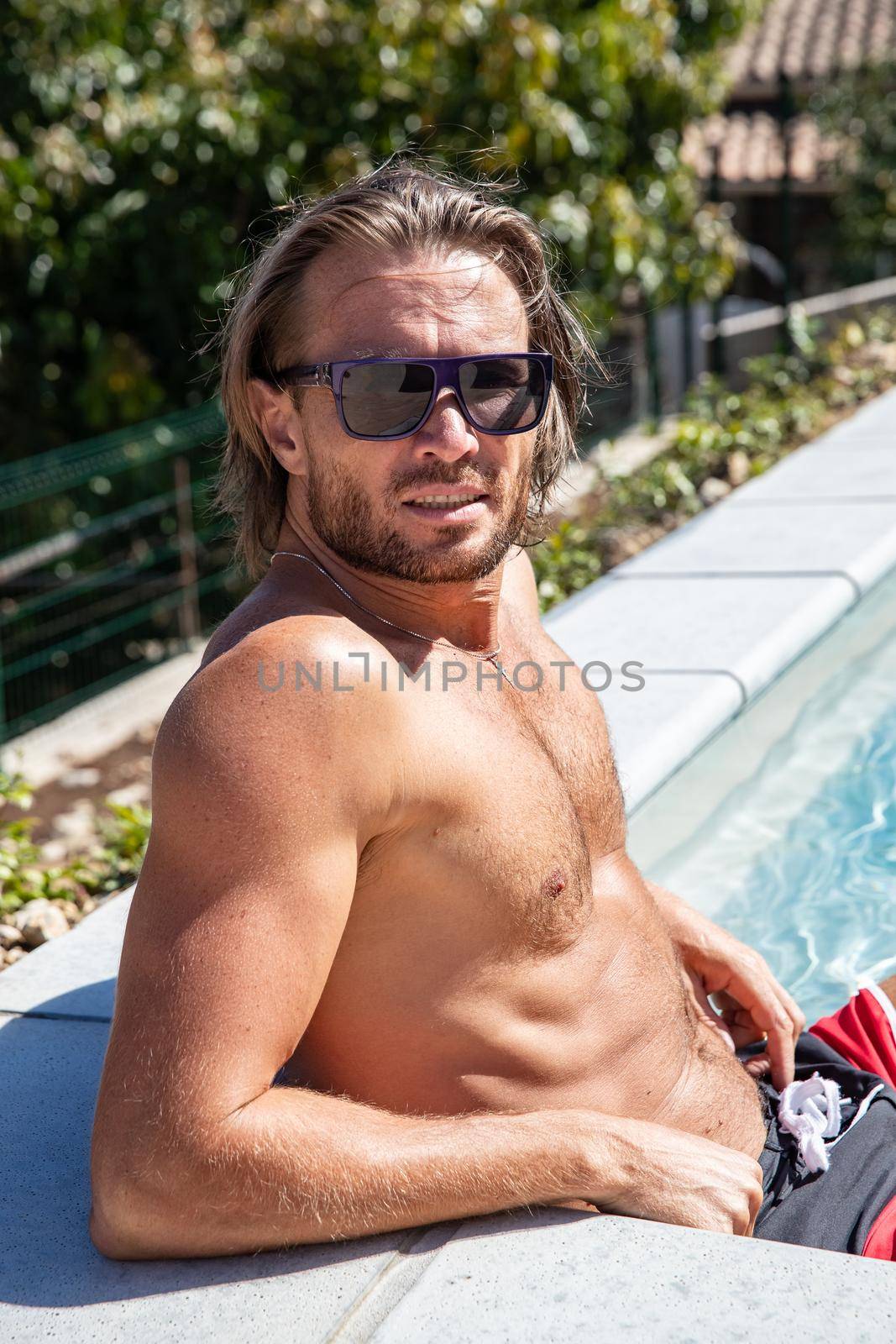 The handsome brutal man in sunglasses with a long hair and naked torso sits near the pool, is looking a camera, a sports suntanned body, sunglasses with a blue frame, he is red blue swimming shorts by vladimirdrozdin