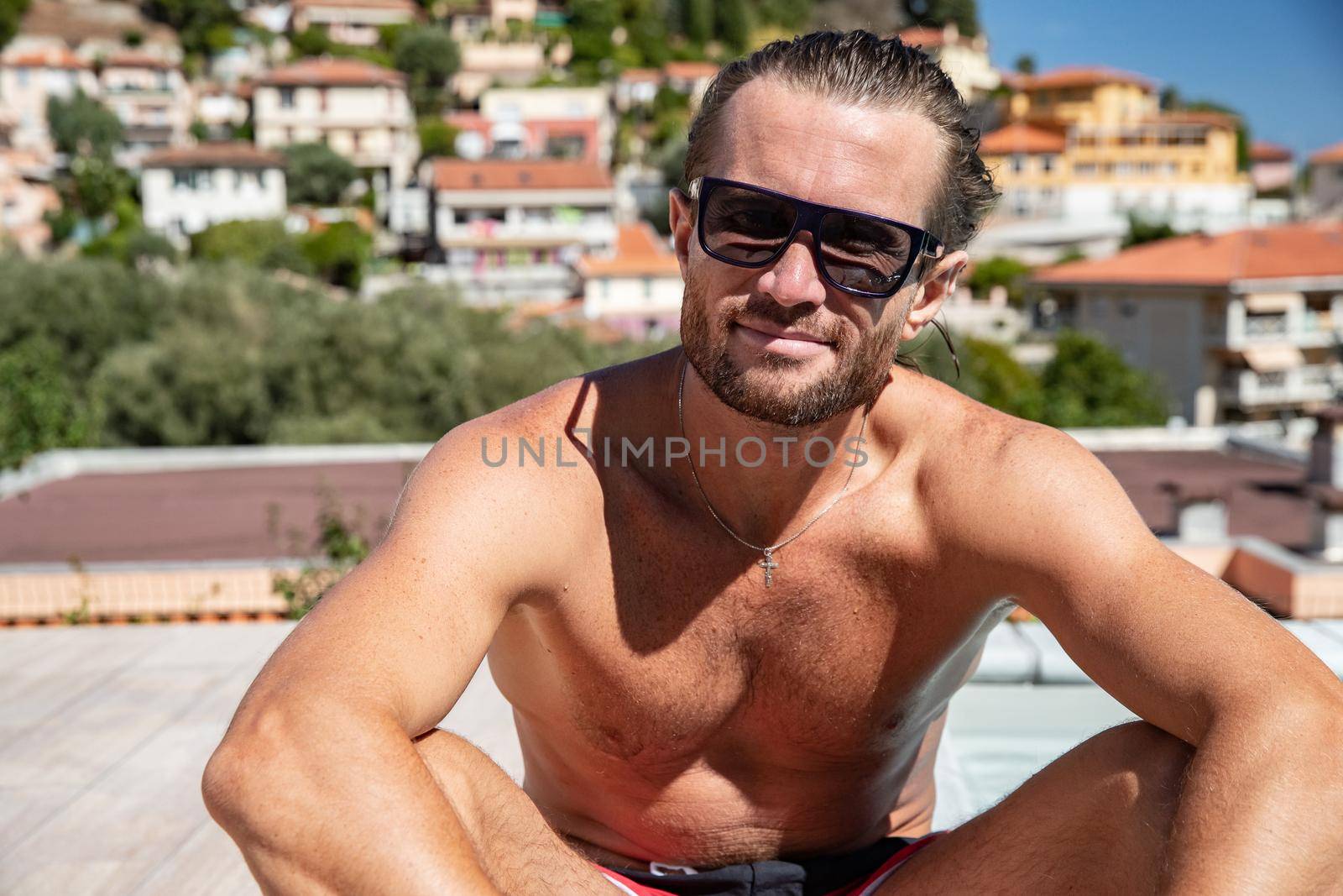 The handsome brutal man in sunglasses with a long hair and naked torso sits near the pool, a sports suntanned body, sunglasses with a blue frame, he is red blue swimming shorts, sunny day