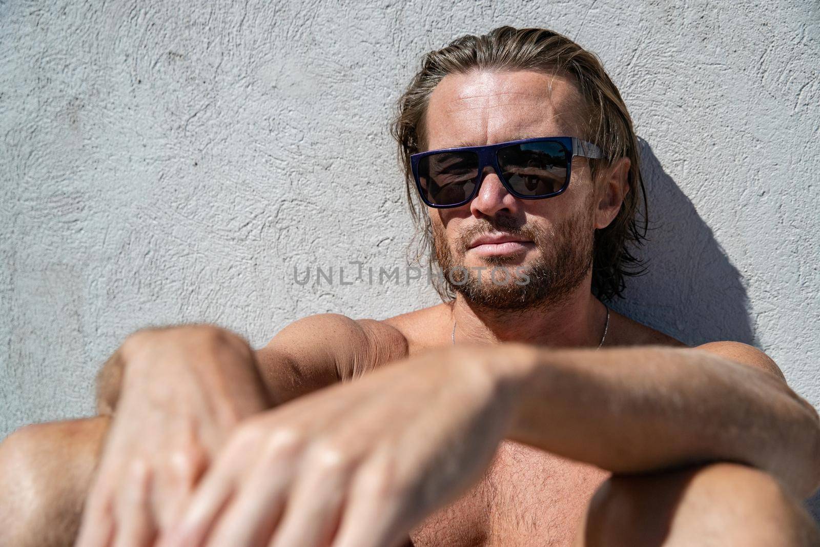 The handsome brutal man in sunglasses with a long hair and naked torso sits near the pool, is looking a camera, a sports suntanned body, sunglasses with a blue frame, he is red blue swimming shorts by vladimirdrozdin