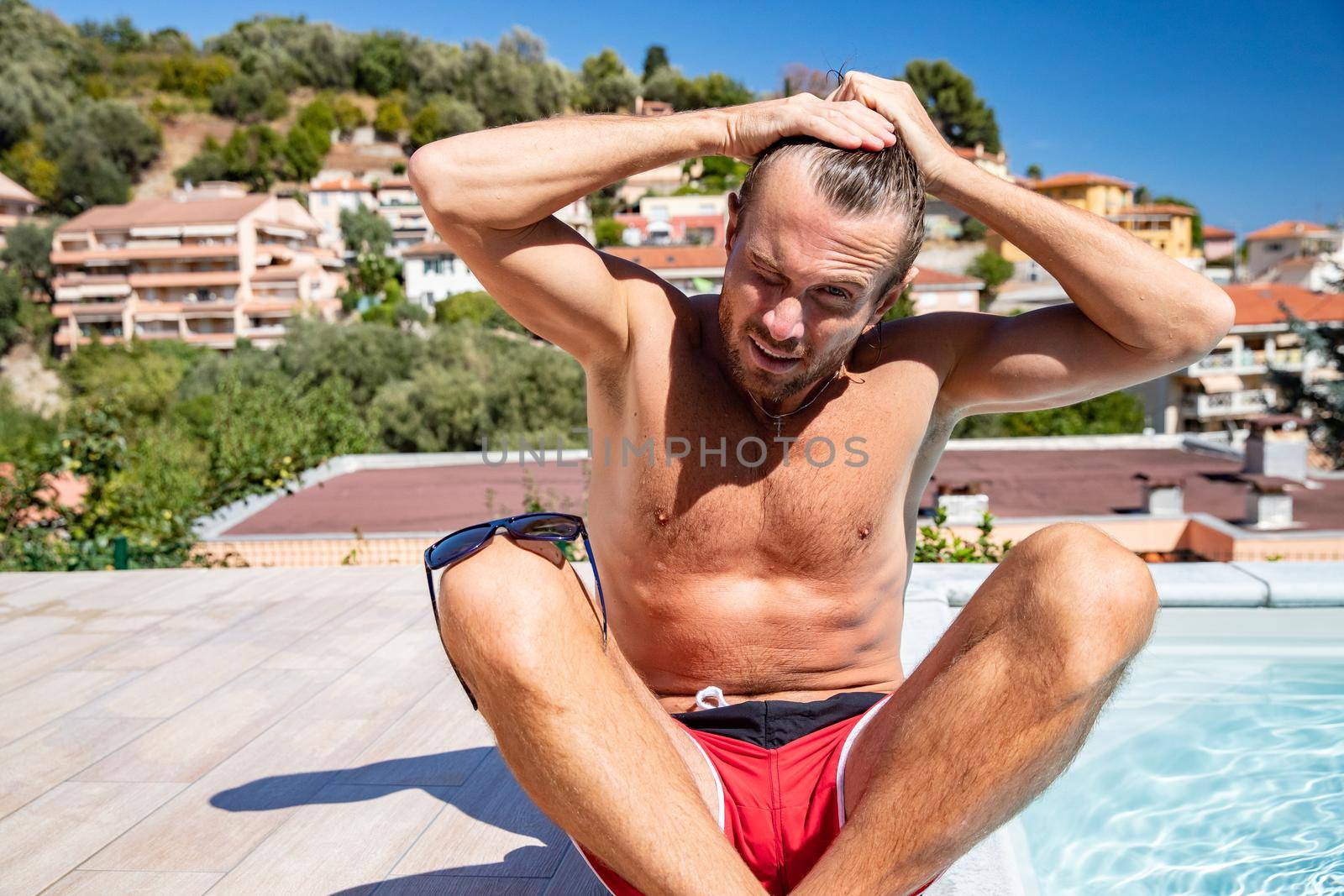 The handsome brutal man in sunglasses with a long hair and naked torso sits near the pool, a sports suntanned body, sunglasses with a blue frame, he is red blue swimming shorts, sunny day
