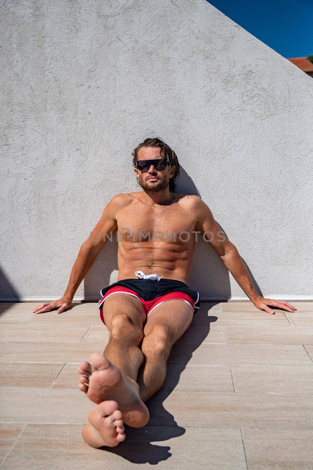 The handsome brutal man in sunglasses with a long hair and naked torso sits near the pool, a sports suntanned body, sunglasses with a blue frame, he is red blue swimming shorts, sunny day