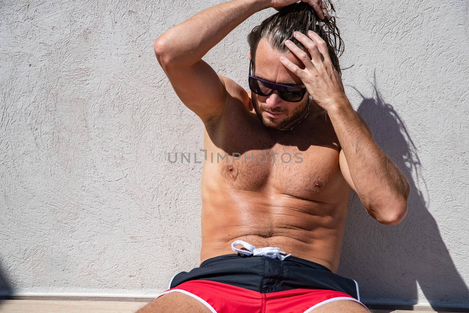 The handsome brutal man in sunglasses with a long hair and naked torso sits near the pool, a sports suntanned body, sunglasses with a blue frame, he is red blue swimming shorts, sunny day