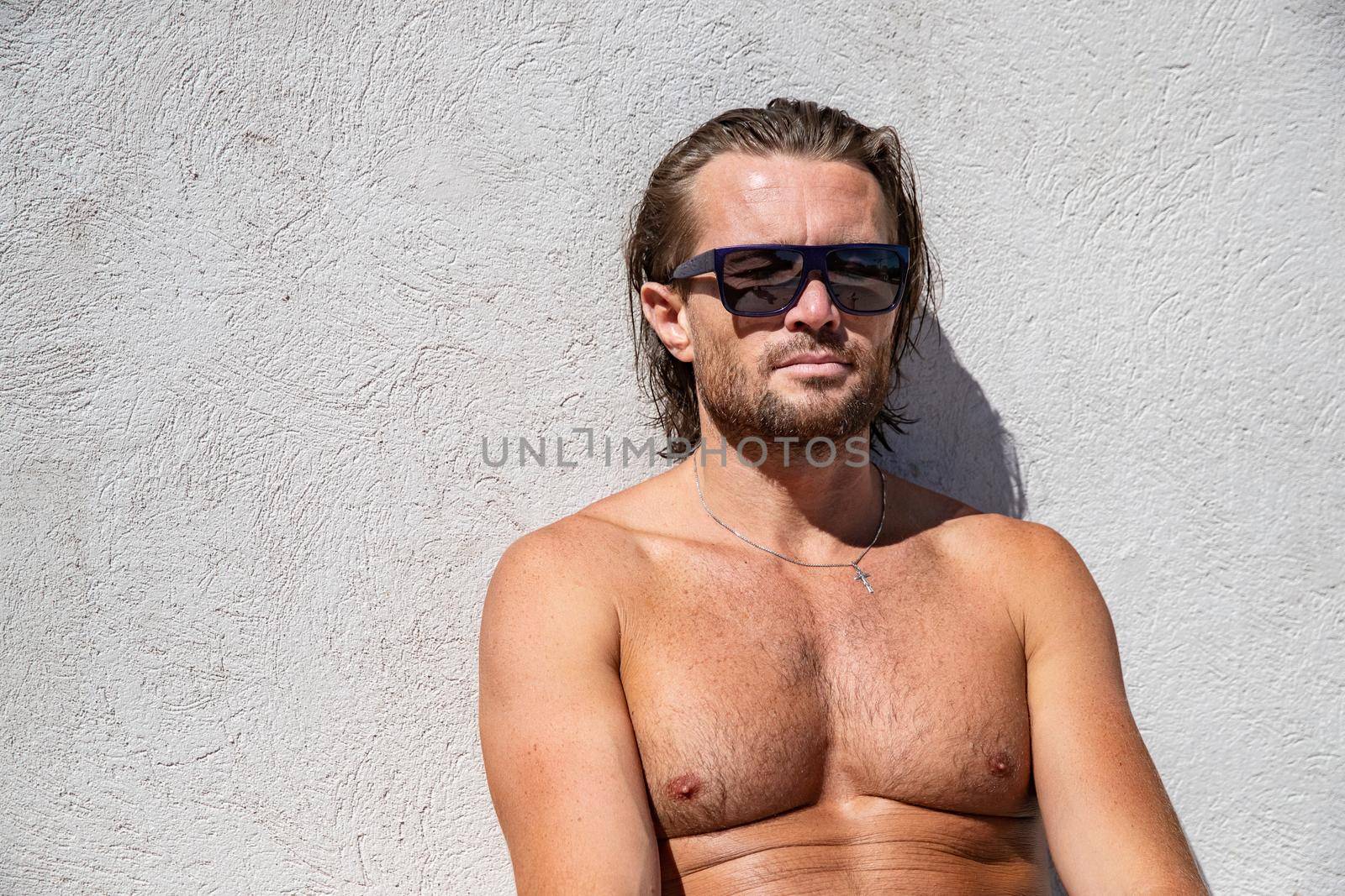 The handsome brutal man in sunglasses with a long hair and naked torso sits near the pool, eyes is closed, a sports suntanned body, sunglasses with a blue frame, he is red blue swimming shorts by vladimirdrozdin