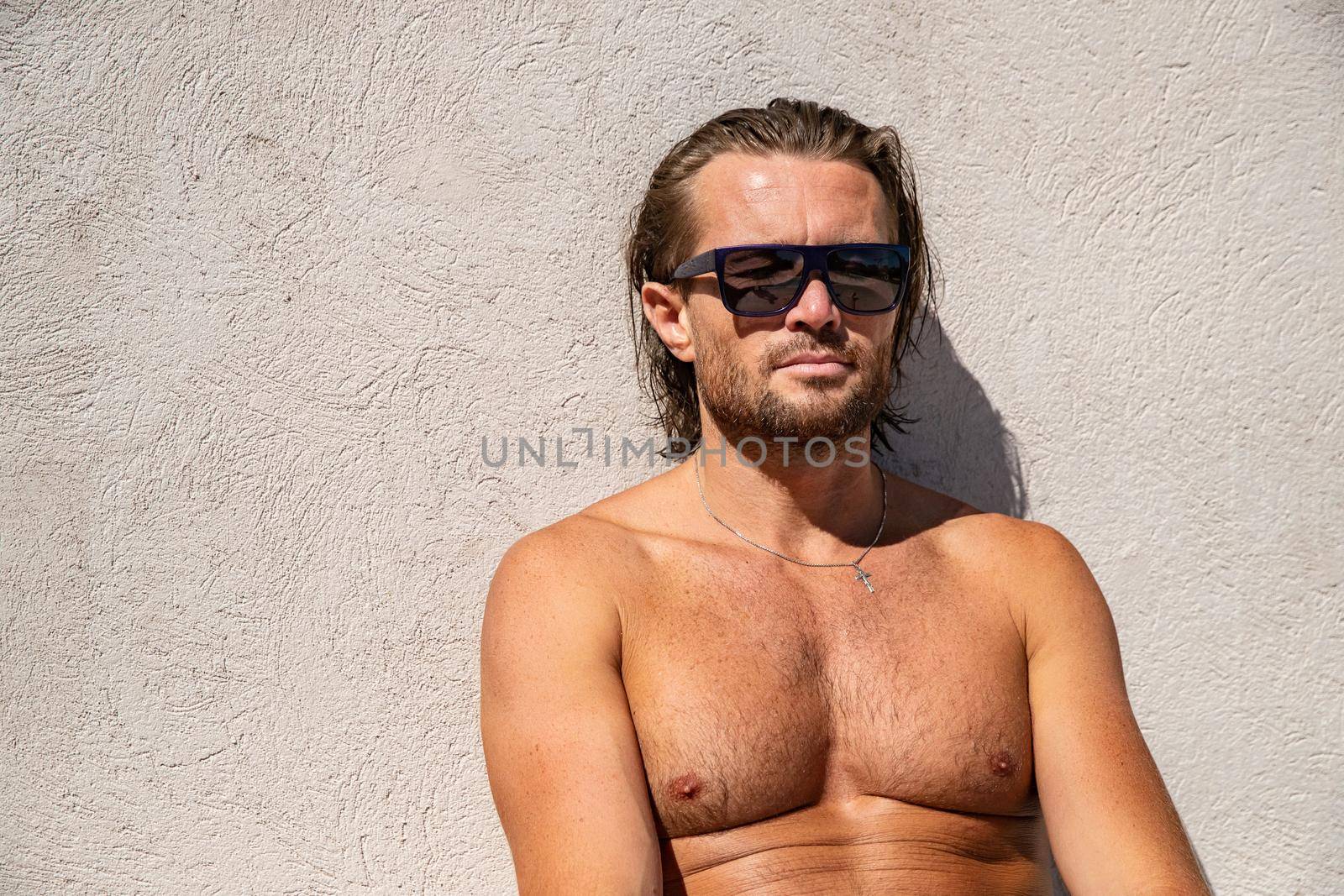 The handsome brutal man in sunglasses with a long hair and naked torso sits near the pool, a sports suntanned body, sunglasses with a blue frame, he is red blue swimming shorts, sunny day