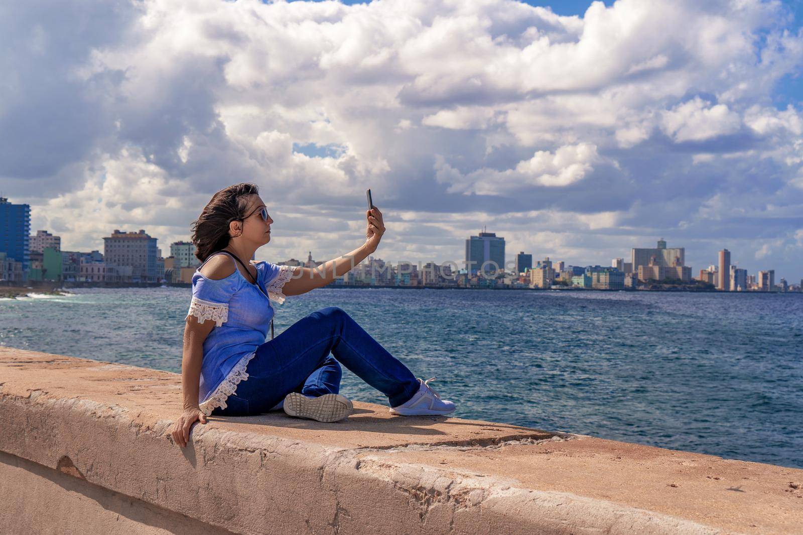 Woman sitting on the Malecon in Havana in front of the sea using a cell phone