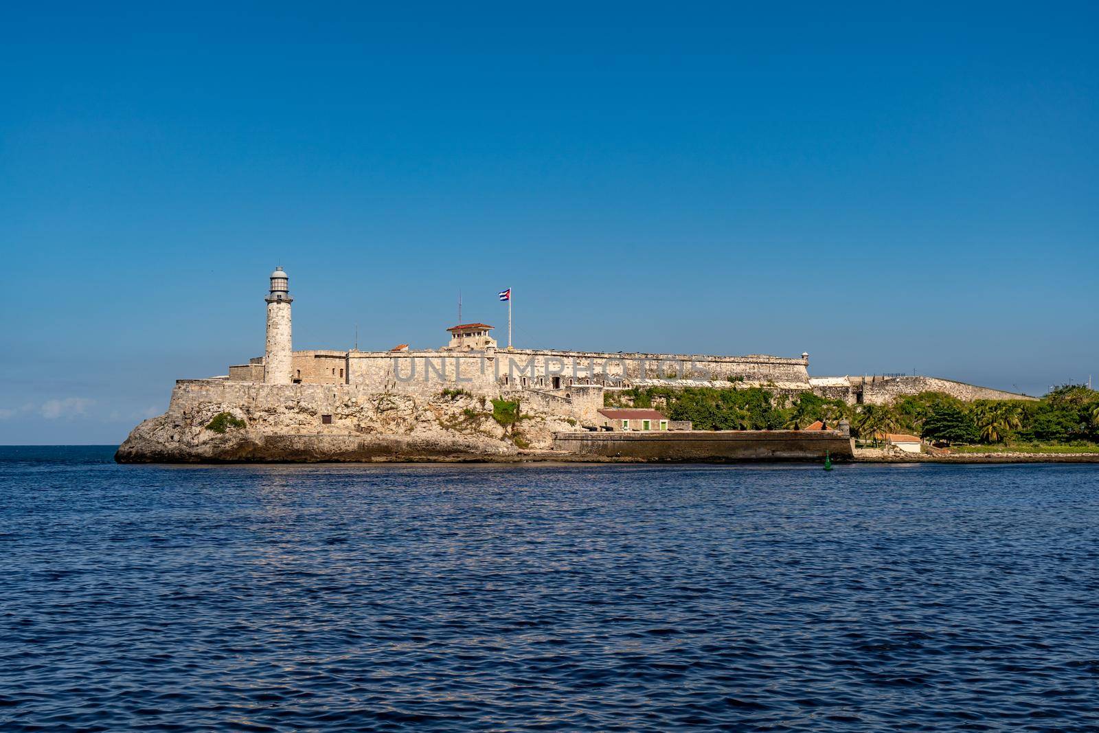 Havana Cuba. November 25, 2020: Horizontal photo of the Morro de la Habana fortress and the lighthouse surrounded by the sea at the entrance to the bay of Havana. In the center the Cuban flag waving on a pole. Famous and tourist place