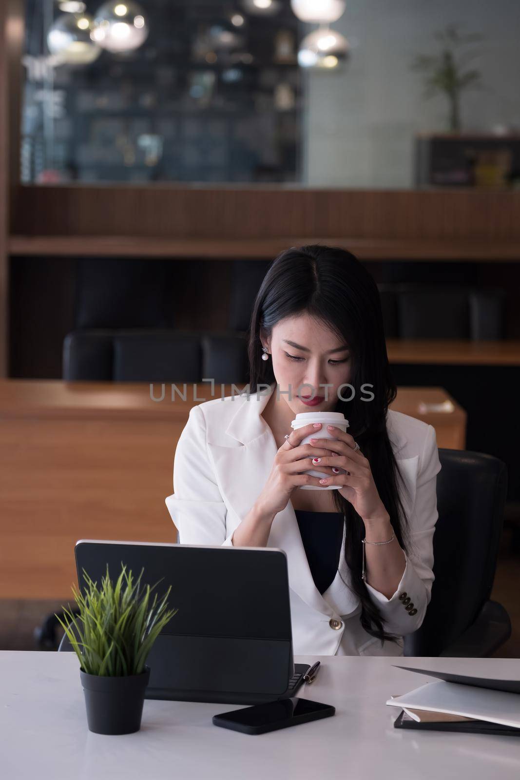 Portrait of business or accountant sitting at desk in modern office with interior drinking hot beverage holding cup in hands while video conference meeting.