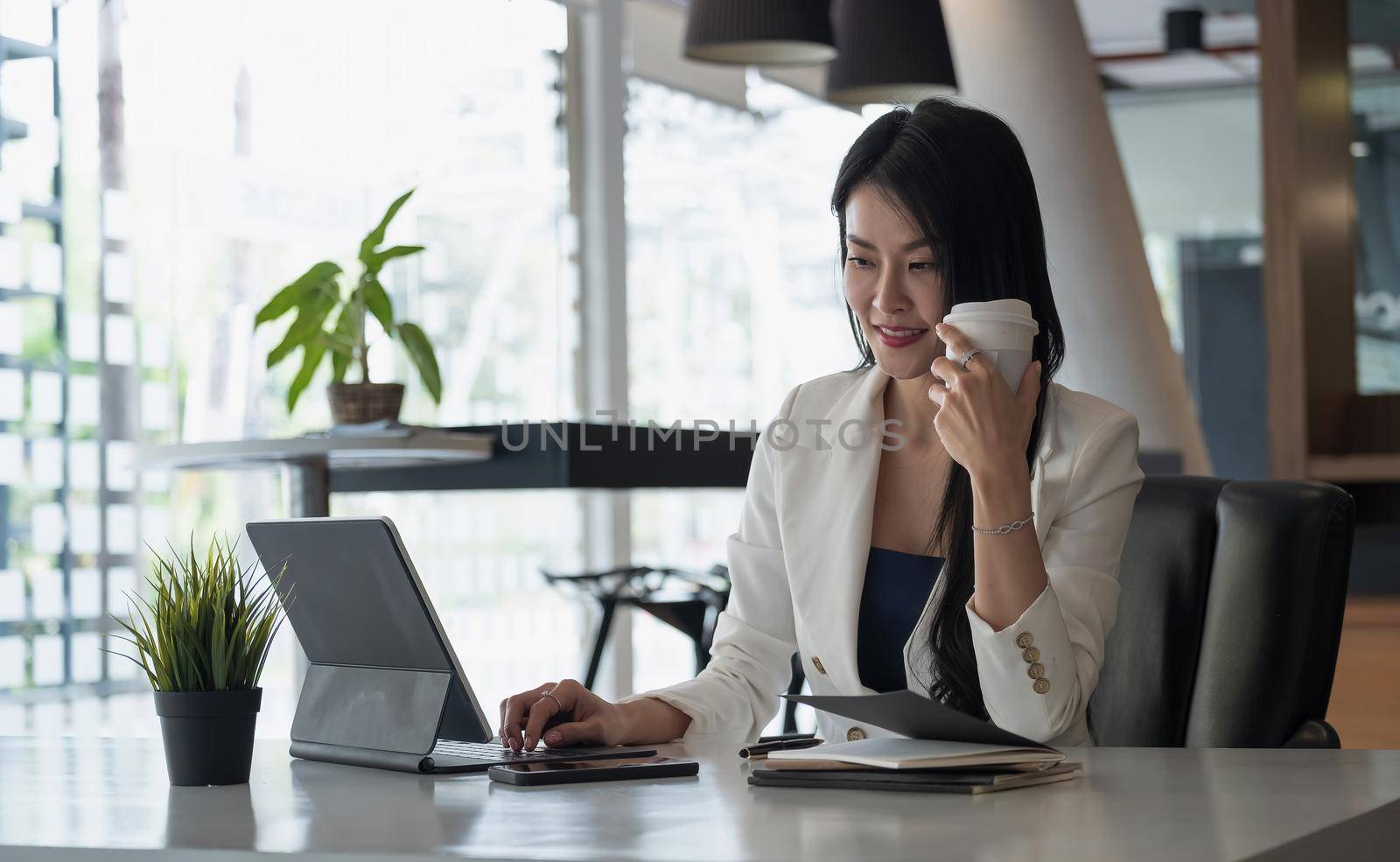 Portrait of business or accountant sitting at desk in modern office with interior drinking hot beverage holding cup in hands while video conference meeting.