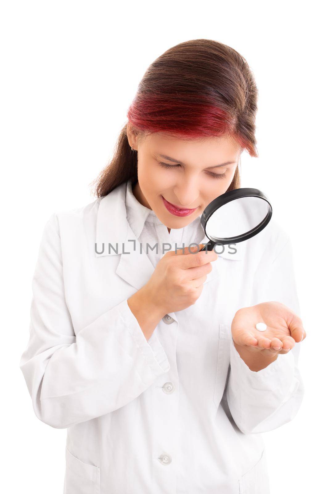 Beautiful smiling young doctor in white lab coat examining a white pill though a magnifying glass, isolated on white background. Scientist with magnifying glass examining pill. Medication scrutiny concept.