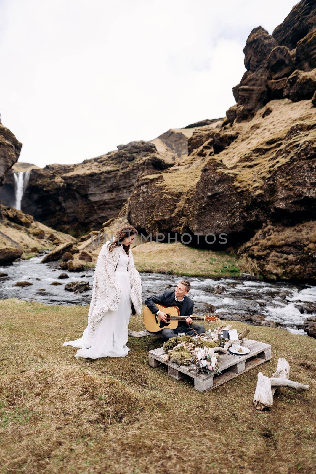 Destination Iceland wedding, near Kvernufoss waterfall. The groom plays the guitar sitting on the grass at an impromptu wedding table, the bride in a white dress and a woolen blanket - dancing nearby by Nadtochiy