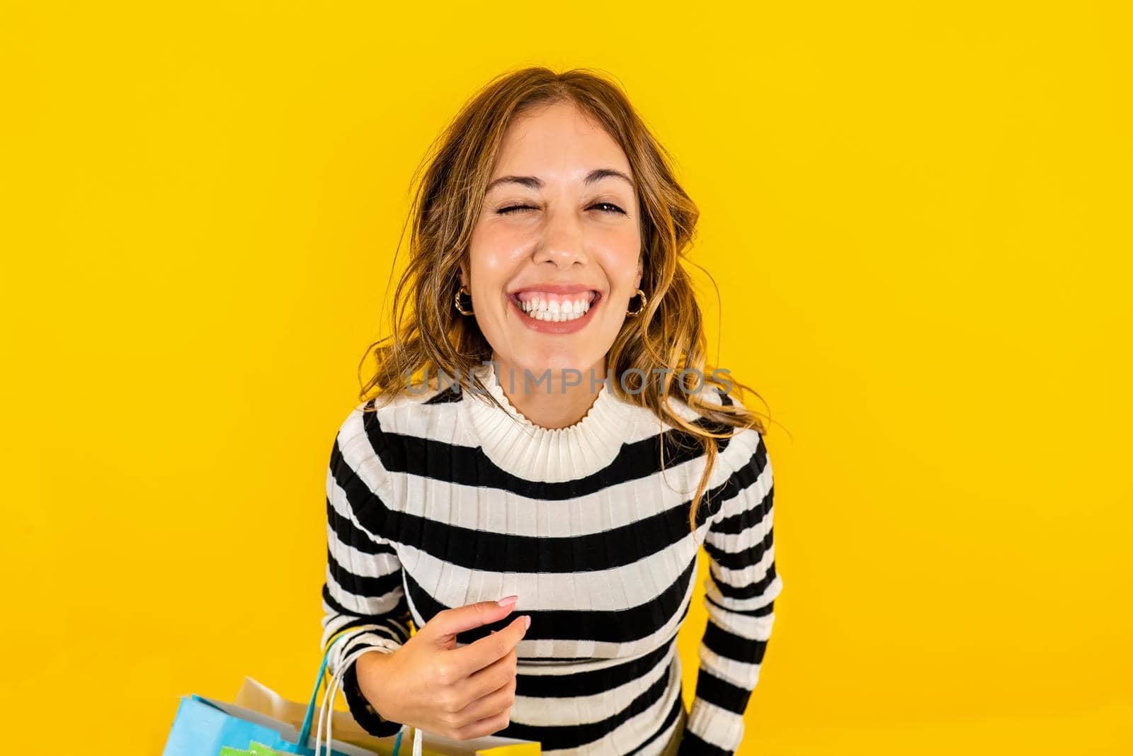 Young beautiful Caucasian woman laughs hard and winking as she looks at the camera happy with her online purchases. Studio shot of happy model holding shopping bags on yellow background for copy space by robbyfontanesi