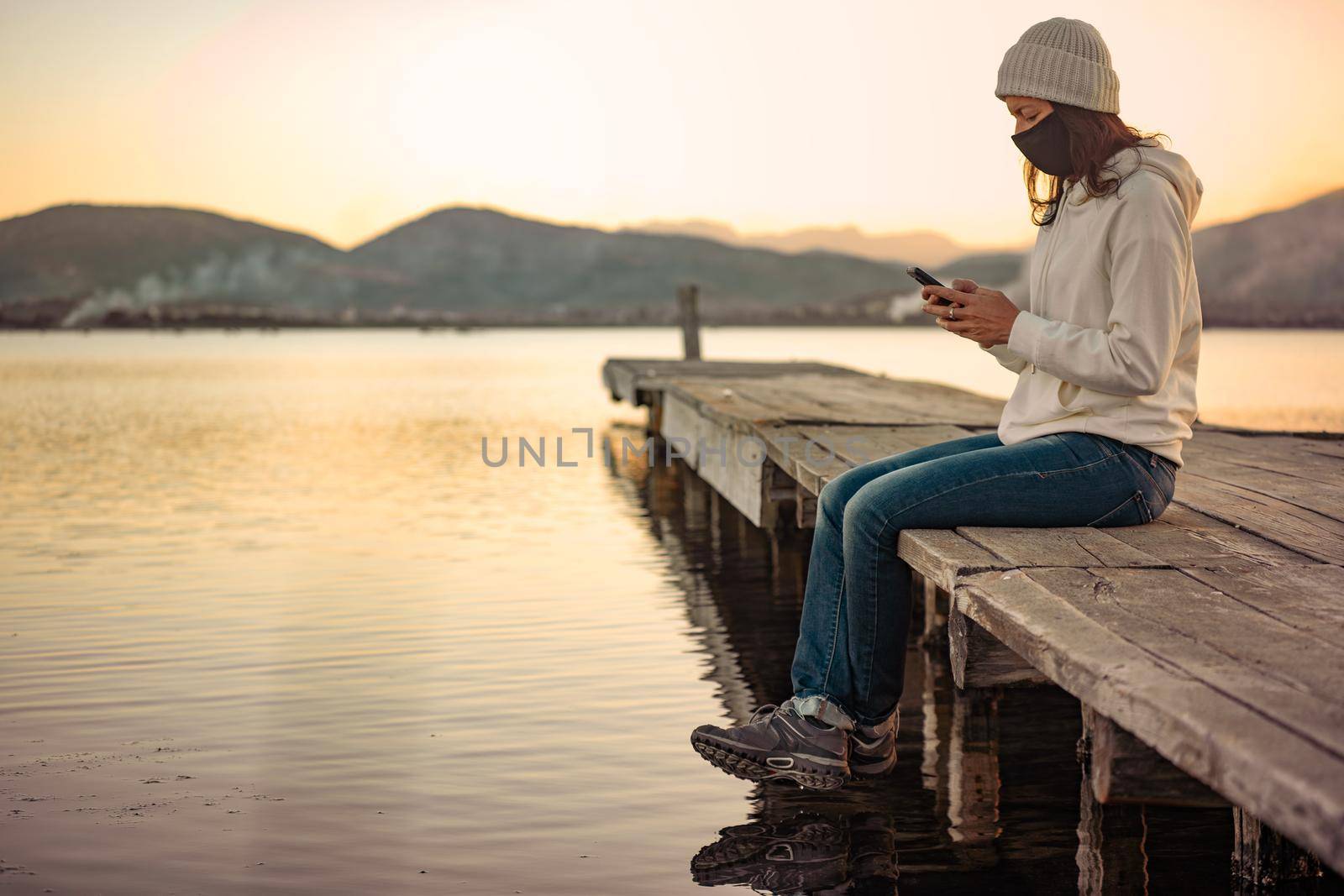 Lonely woman uses her smartphone to follow the latest news on Covid-19 sitting on a pier with protective mask at sunset or sunrise. New normal activity with internet mobile technology with Coronavirus