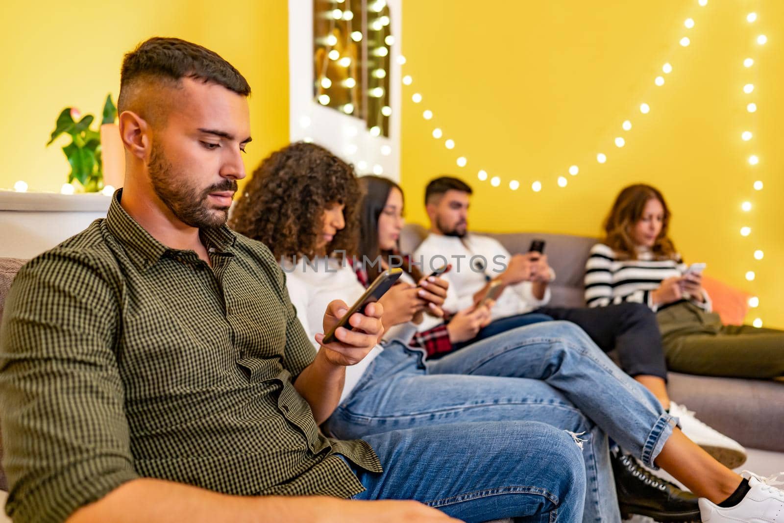 Group of serious young student friends at home sitting on the couch spend time using smartphones without real interaction between them. New social and human habits due to internet mobile technology