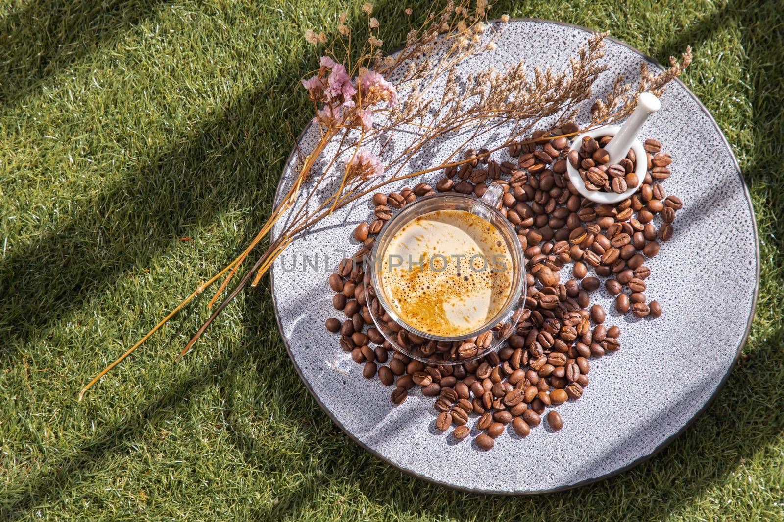 Coffee style, Black coffee in glass cup with coffee beans roating and dried flower on Ceramic plate at lawn in the morning. Top view.