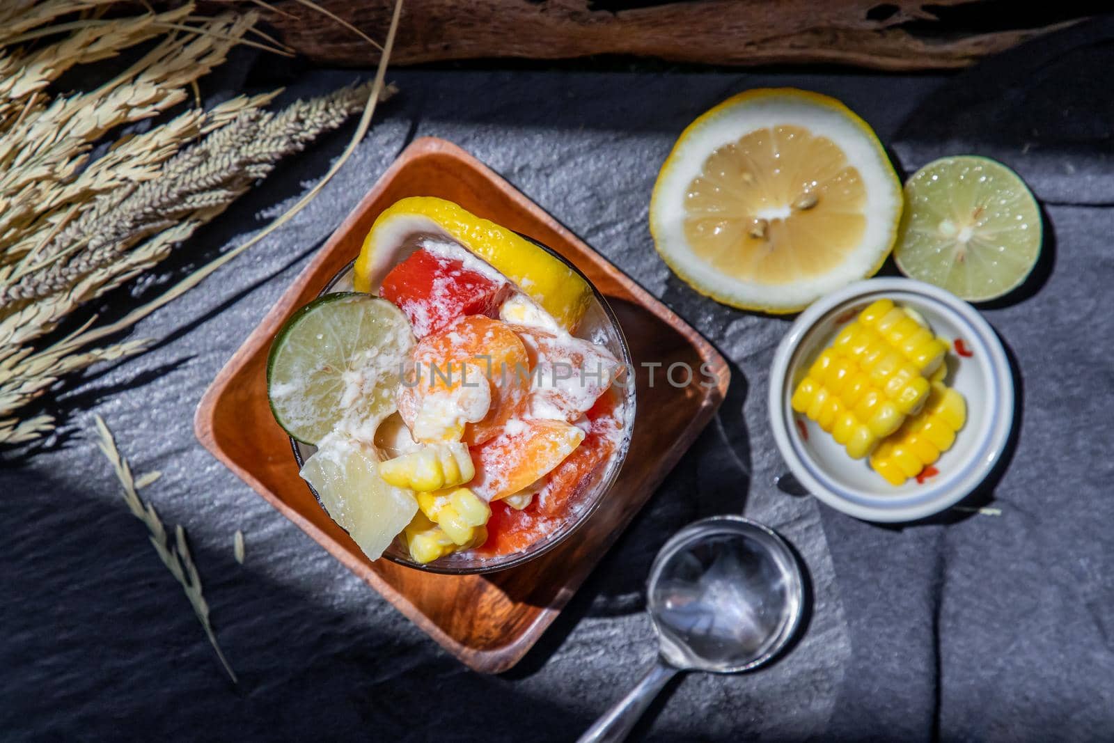 Tropical fresh fruit salad with milk and oatmeal in a glass cup served with corn on dark background. Sweet healthy breakfast, Healthy vegan meal. Top view, Selective focus.