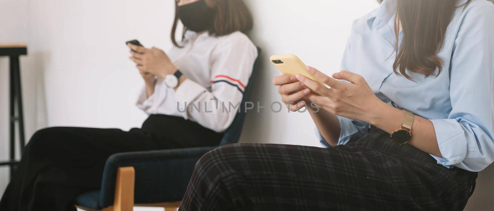Two Asian young woman chatting in smartphone and wearing mask sitting distance protect from COVID-19 viruses for social distancing for infection risk. by nateemee